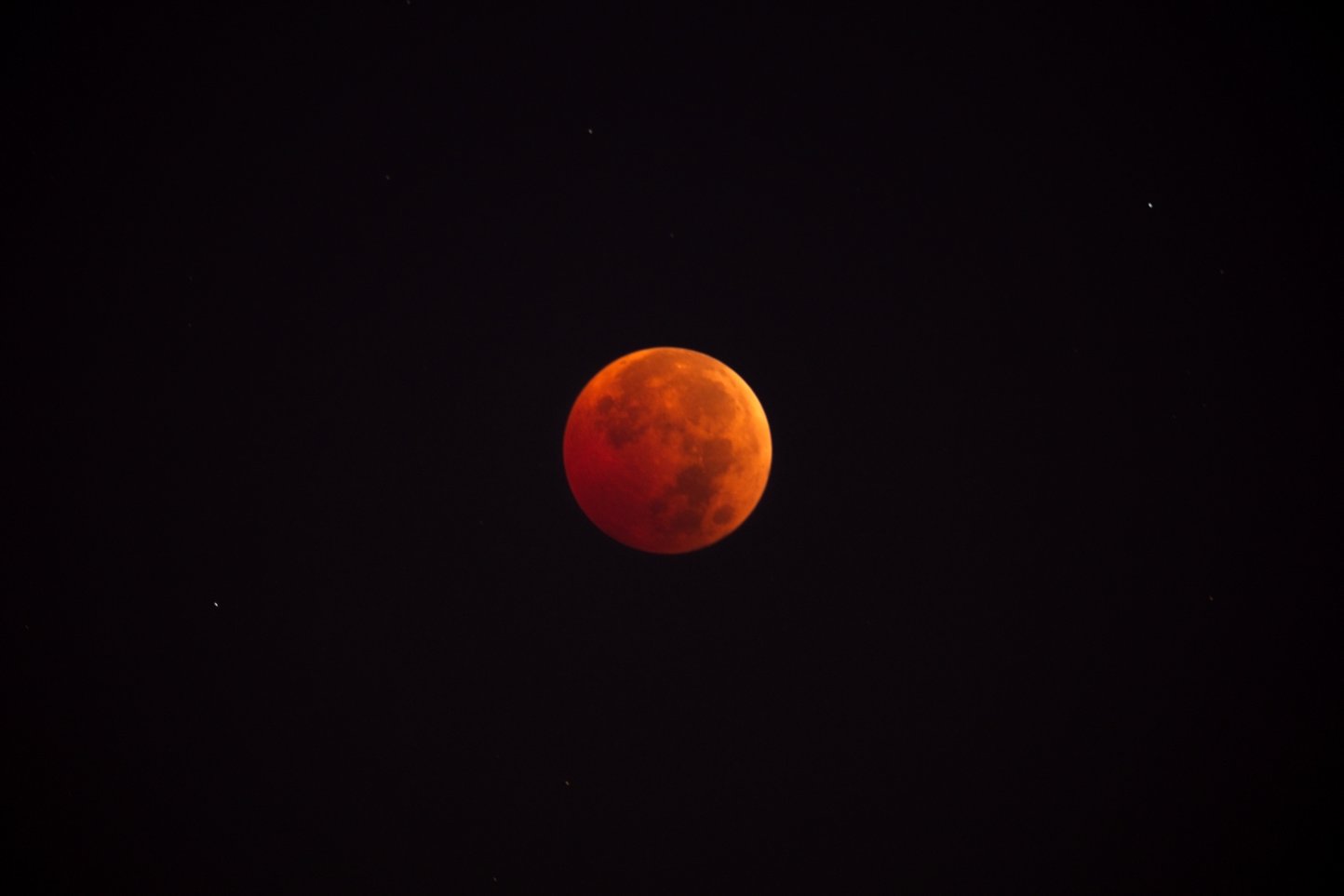 A blood moon glowing in deep red-orange during a total lunar eclipse, set against a dark night sky with a few faint stars scattered around.