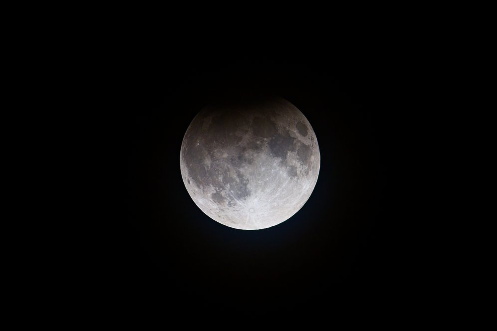 A close-up view of the full Moon centered against a pitch-black background. The lunar surface displays detailed craters and darker maria (ocean-like basins), with a subtle shadow or partial eclipse shadow visible across the upper portion. The lower half is brightly illuminated, highlighting the texture and rugged terrain of the Moon.