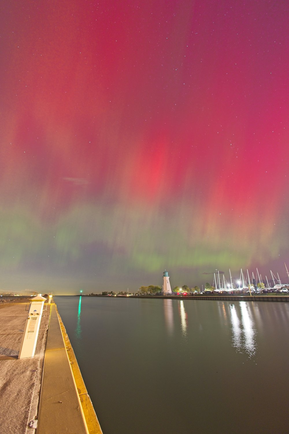 A breathtaking night scene of the aurora borealis (northern lights) painting the sky in vibrant red and green hues. In the foreground, a concrete pier extends toward the water, with a lighthouse standing near a marina filled with docked sailboats. The calm water reflects the glowing lights of the lighthouse, marina, and aurora, while stars are scattered across the colorful night sky.