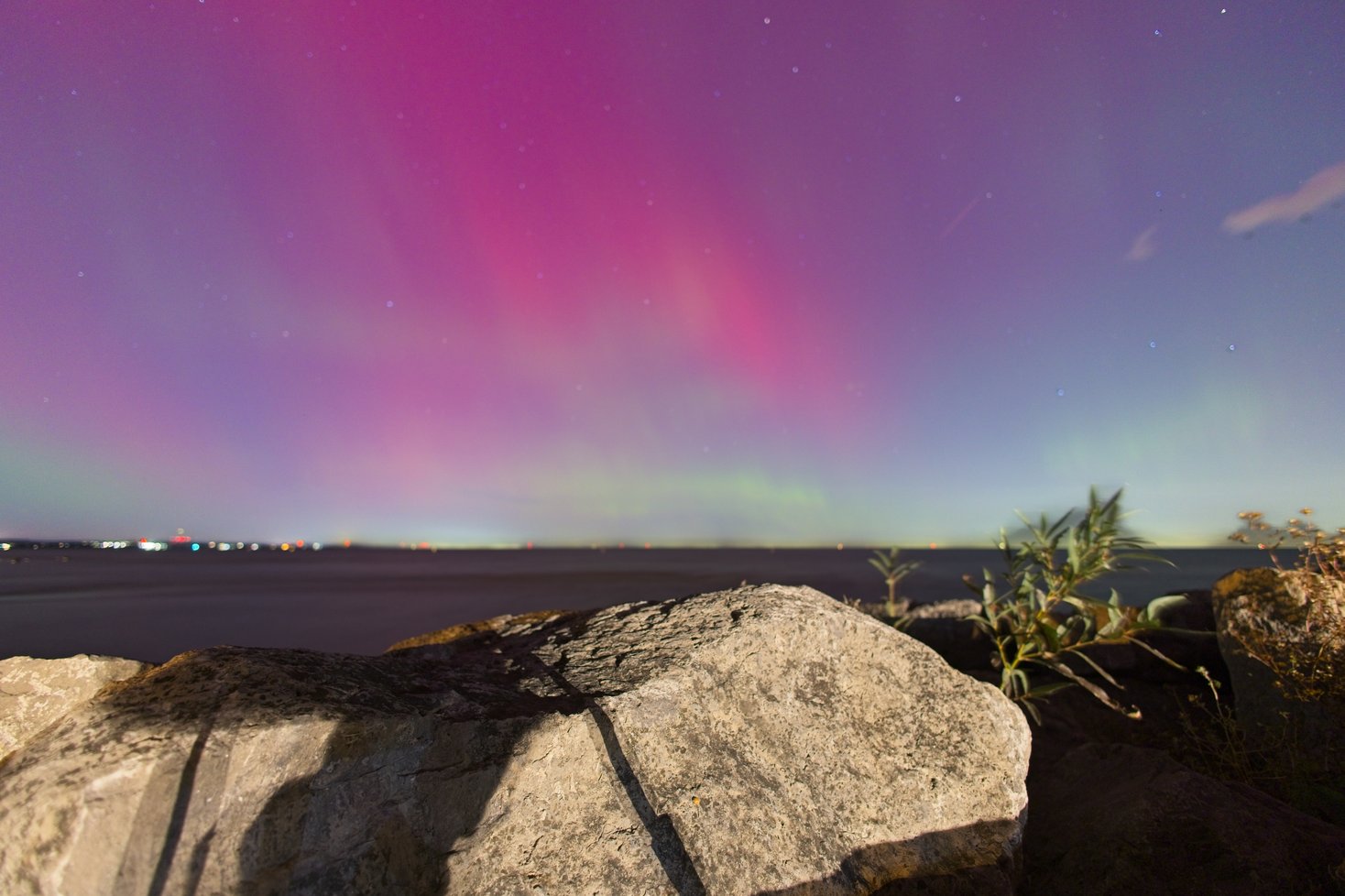 A breathtaking night scene of the aurora borealis (northern lights) painting the sky in vibrant red and green hues. In the foreground, a concrete pier extends toward the water, with a lighthouse standing near a marina filled with docked sailboats. The calm water reflects the glowing lights of the lighthouse, marina, and aurora, while stars are scattered across the colorful night sky.