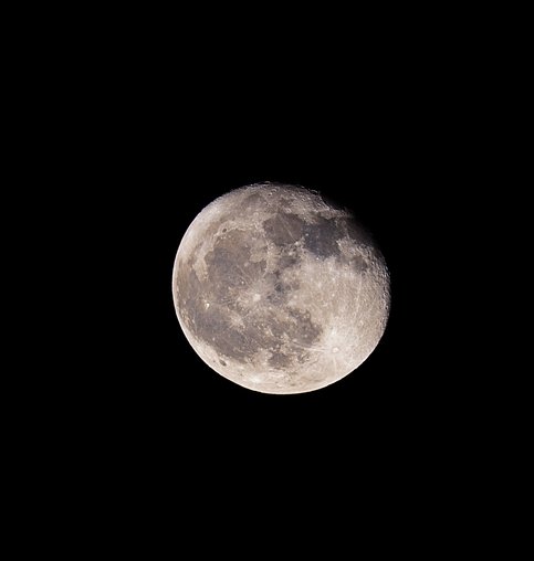 A close-up view of the full Moon centered against a pitch-black background. The lunar surface displays detailed craters and darker maria (ocean-like basins), with a subtle shadow or partial eclipse shadow visible across the upper portion. The lower half is brightly illuminated, highlighting the texture and rugged terrain of the Moon.