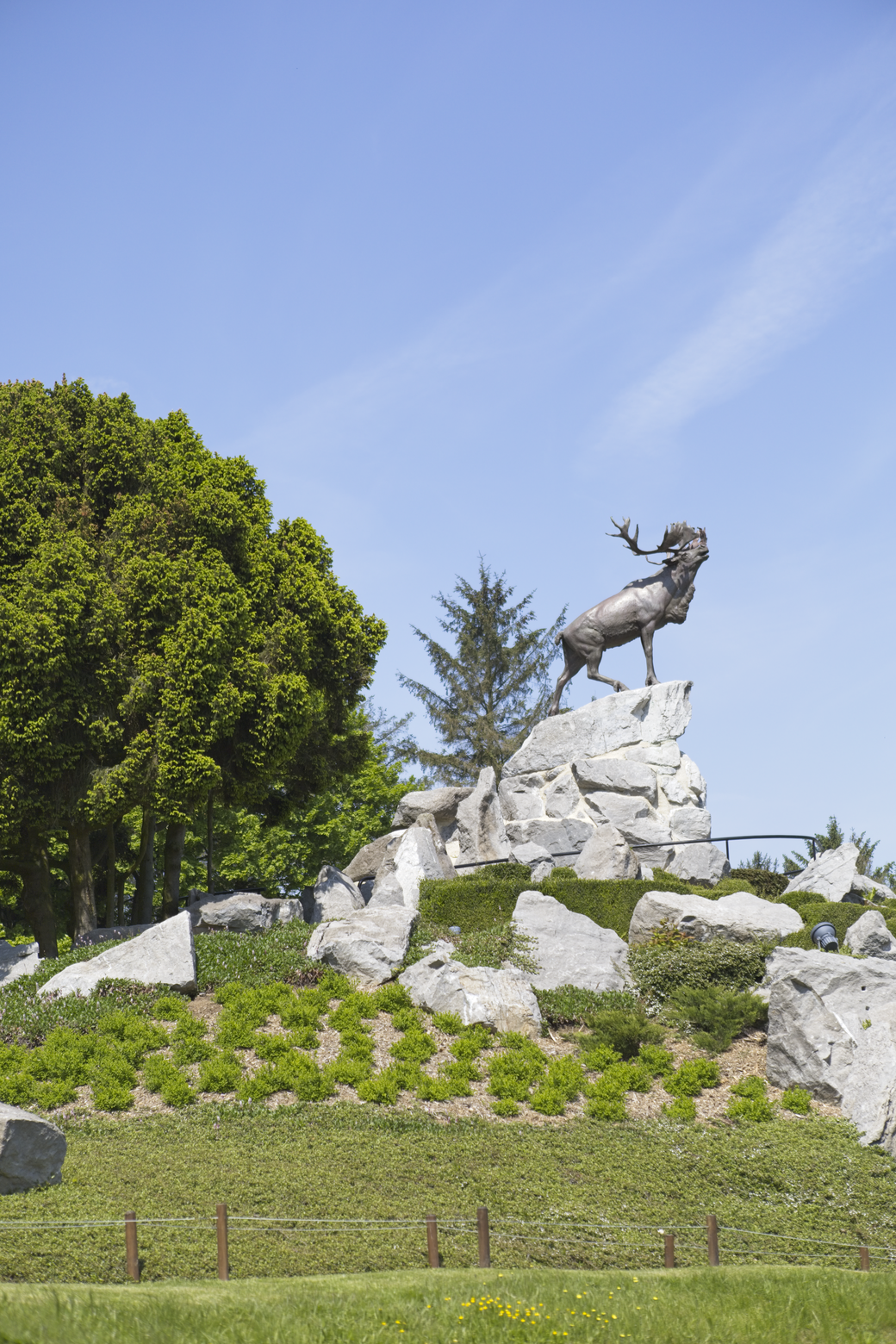 A statue of a stag stands dramatically on large, light-colored rocks in an outdoor park setting. The scene is framed by dense, lush green trees on the left and under a bright blue sky.