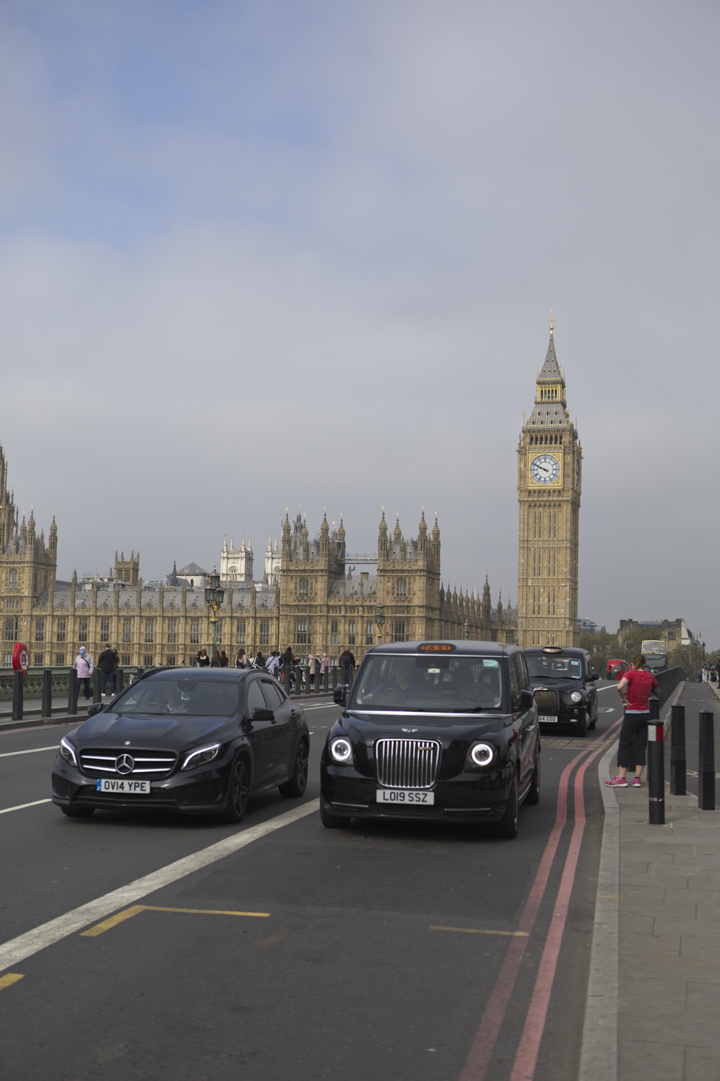 A street view in London featuring the Palace of Westminster and the Elizabeth Tower (Big Ben) prominently in the background under an overcast sky. Several parked or driving vehicles, including a black sedan and a black Mini Cooper, occupy the foreground street.