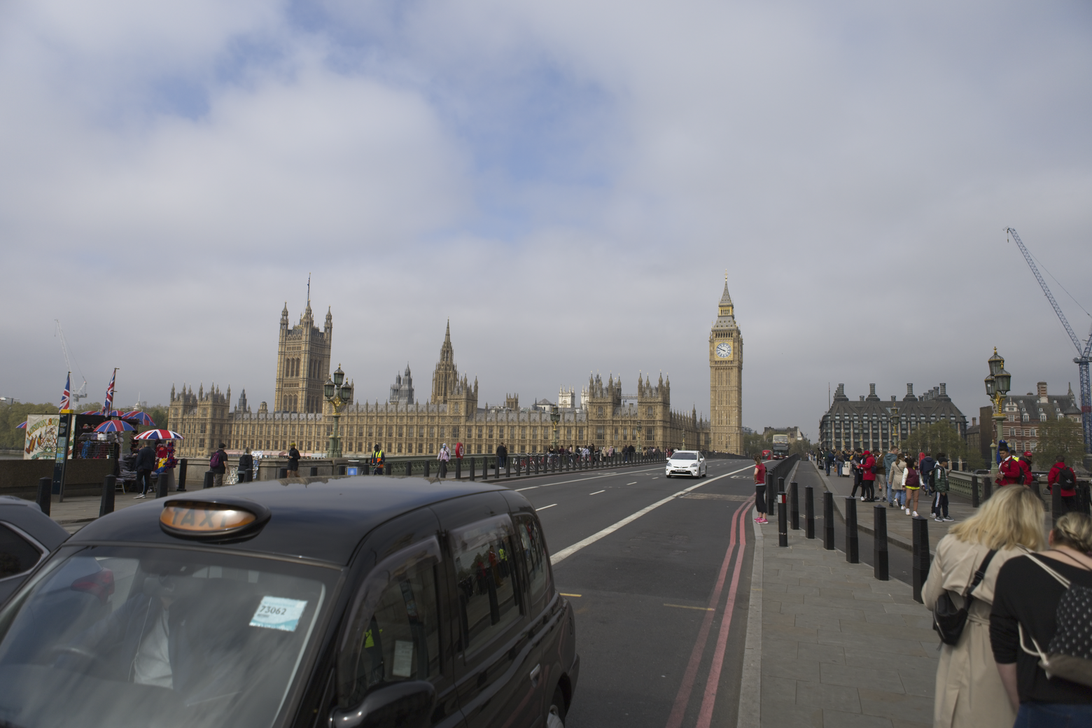 A street-level view of London's historical heart, featuring the Houses of Parliament and the Elizabeth Tower (Big Ben) dominating the background. A wide road extends toward the landmarks, with a large black van in the immediate foreground, and pedestrians visible walking along the sidewalk under an overcast sky.