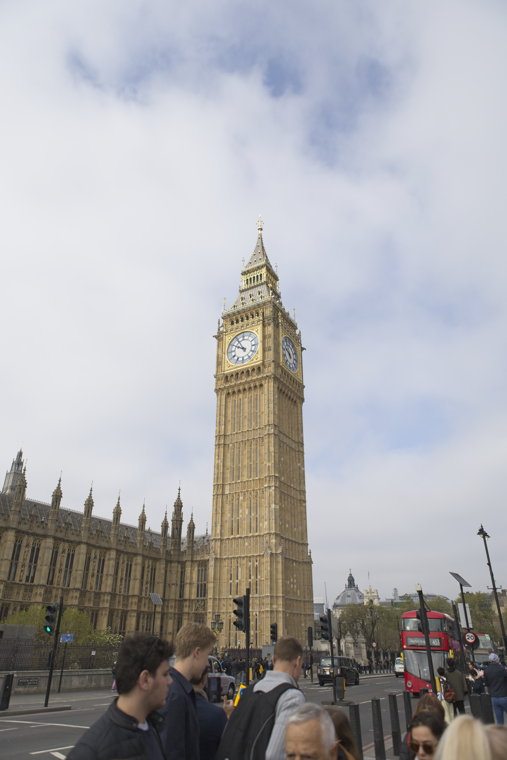 A view of the Houses of Parliament in London, featuring the prominent Elizabeth Tower (Big Ben) and the adjacent Gothic architecture of the Palace of Westminster under a cloudy sky. Several people are visible in the street scene.