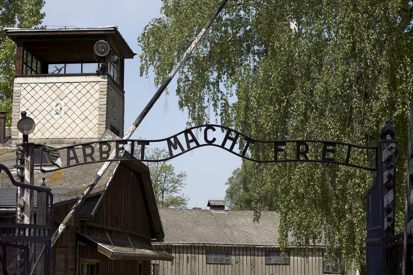 A photo of a weathered entrance archway displaying the German slogan "ARBEIT MACHT FREI" (Work Makes Free), situated in front of aged wooden buildings and framed by dense, vibrant green foliage.
