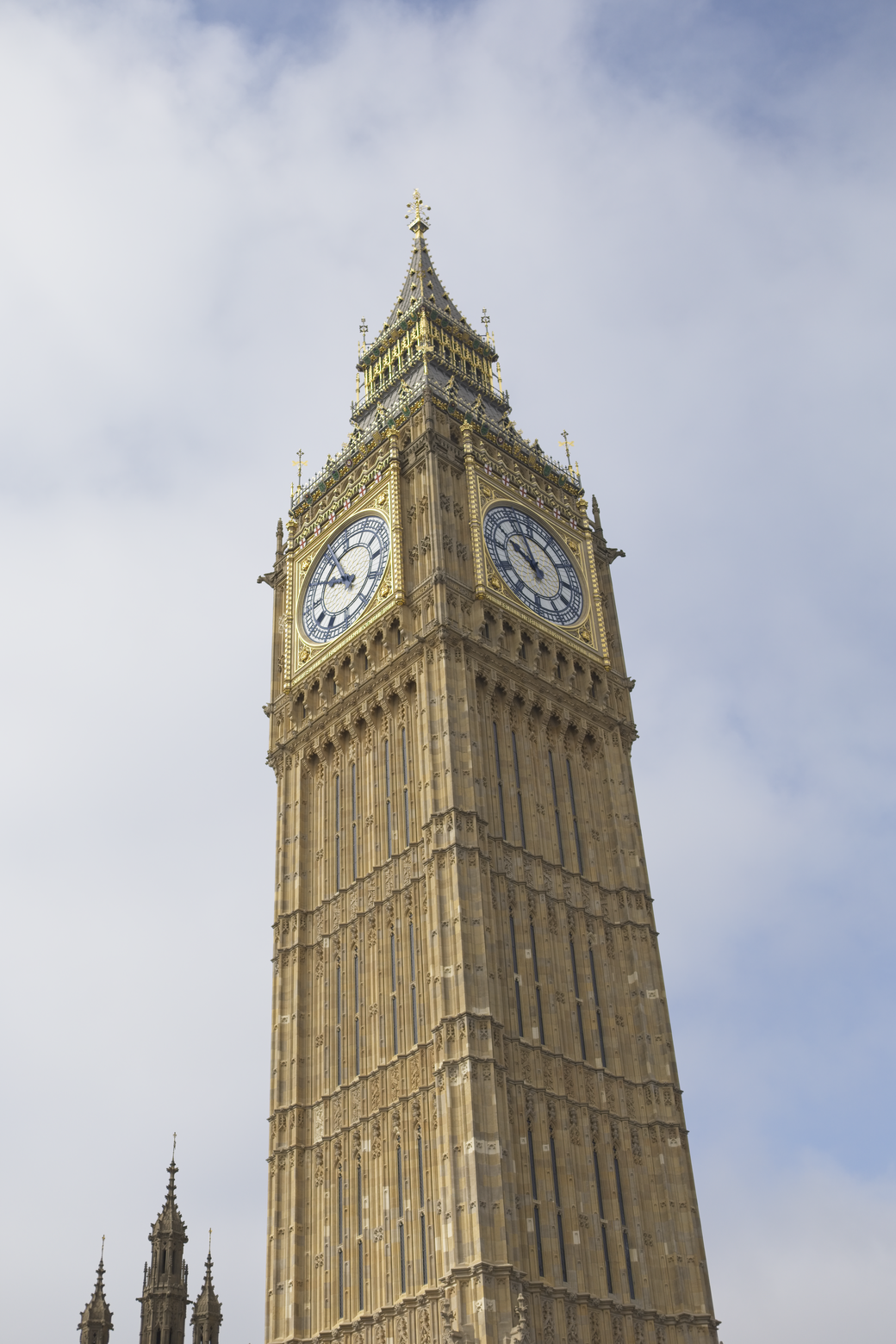 A towering, ornate clock tower, resembling Big Ben/Elizabeth Tower, is viewed from a low angle against a bright, cloudy sky. The massive, detailed structure features visible clock faces near its apex and displays classical Victorian Gothic architecture.
