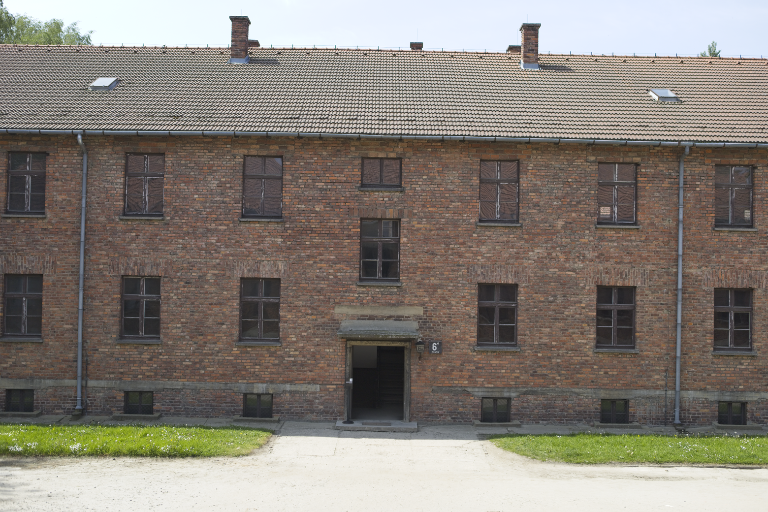 The image captures a large, two-story institutional building constructed of red brick, topped with a tiled roof and featuring several visible chimneys. The facade is symmetrical, with numerous rectangular windows, and is approached via a central pathway leading to a recessed entrance, set against a foreground of green lawn and gravel.