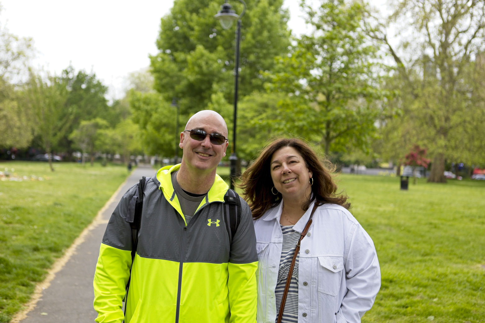 A man and a woman are standing together in a sunny, green park setting. The man is positioned in the foreground wearing a bright yellow and gray athletic jacket, and the woman is beside him wearing a white jacket. They are posing for a casual outdoor portrait with lush trees and grass visible in the background.