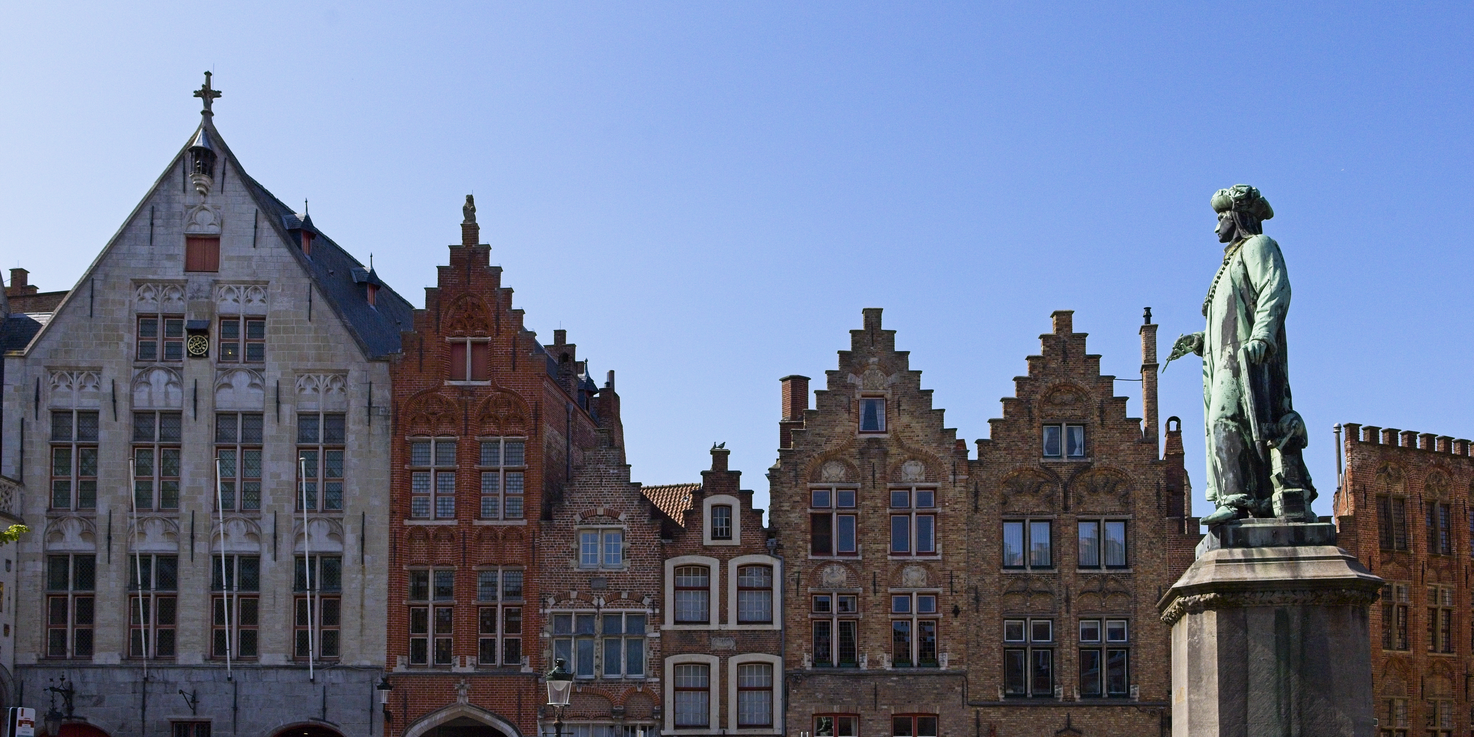 A sunny view of historical European architecture, featuring a row of gabled, multi-story brick buildings under a clear blue sky. On the right side, a prominent statue of a man stands on a raised pedestal, contrasting with the ancient masonry backdrop.