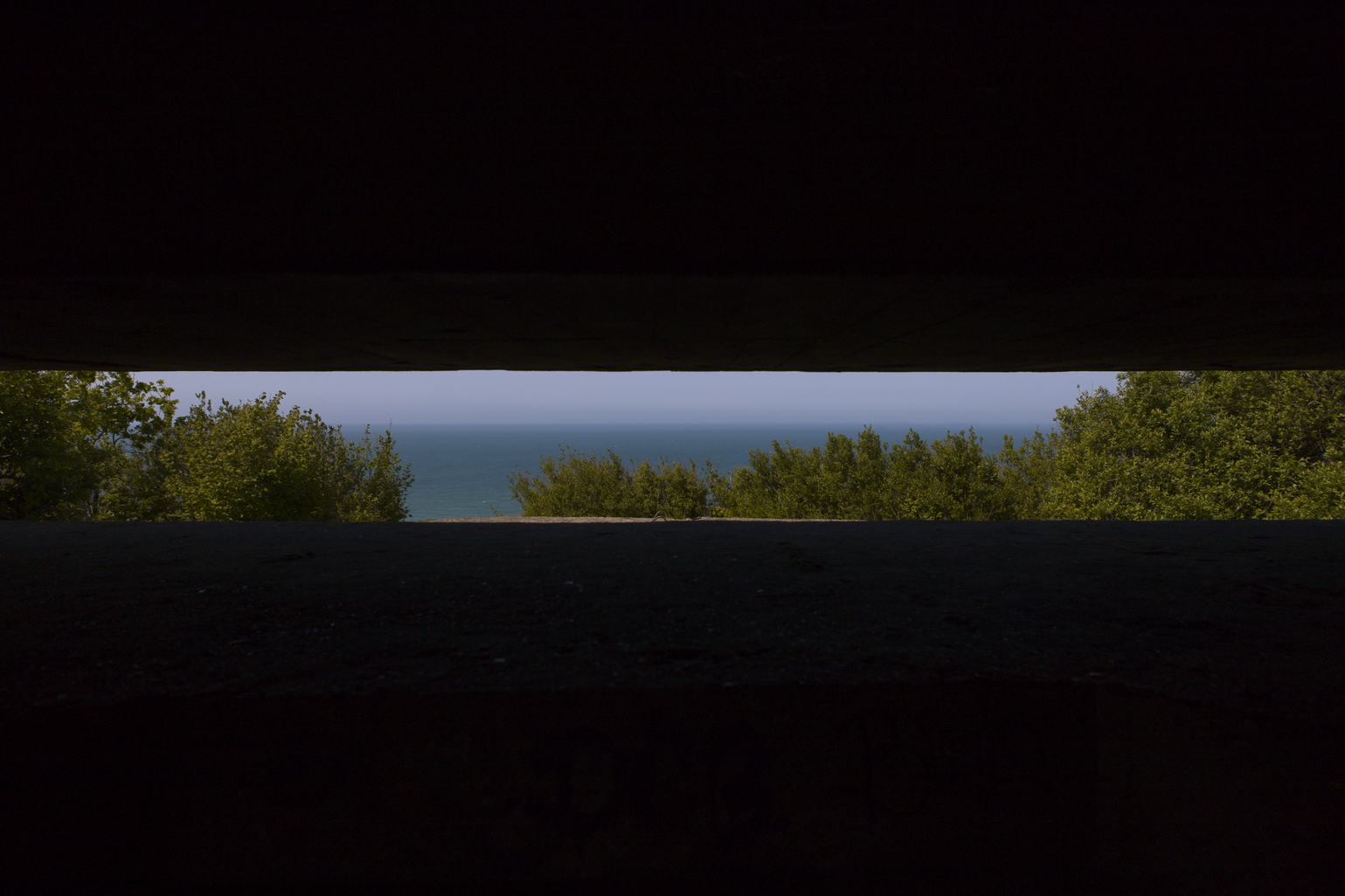 A high-contrast photograph showing a view looking out from under a dark overhang or shadow. The scene features a dark, solid foreground (like a road) framed by green foliage, offering a glimpse of a bright blue expanse (sky or water) in the distance.