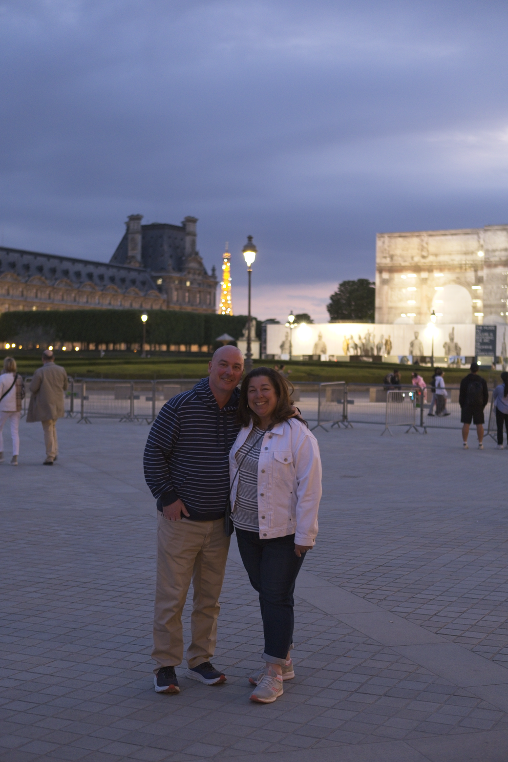 A couple stands in a large, paved plaza at dusk, with impressive, illuminated classical architecture and buildings visible in the background, suggesting a romantic scene at a major European city landmark.