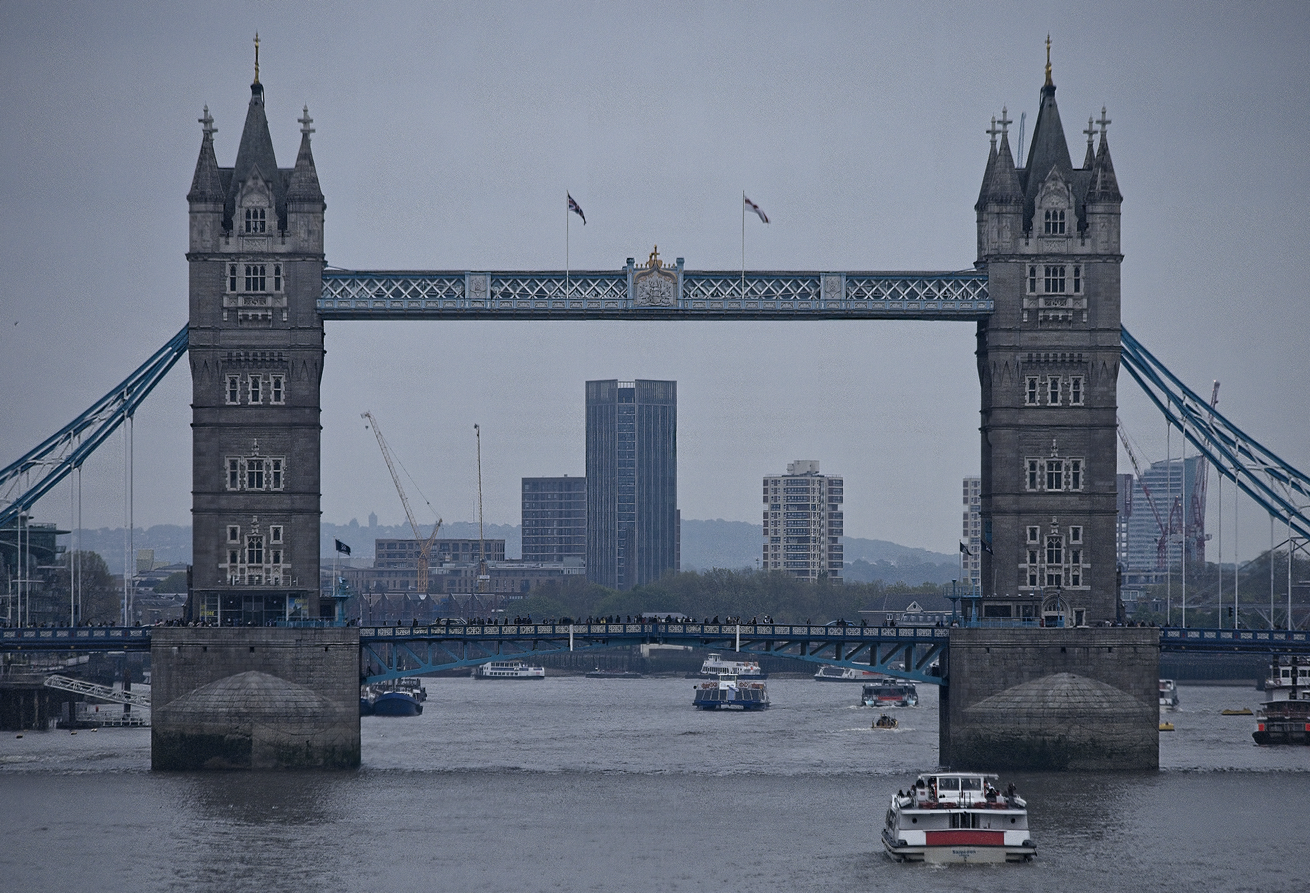 A view of the historical London Bridge, characterized by its imposing twin towers, crossing the River Thames under a heavily overcast sky. The scene captures an urban waterway, with various boats traversing the river beneath the bridge's span, framed against a backdrop of modern city skyscrapers.