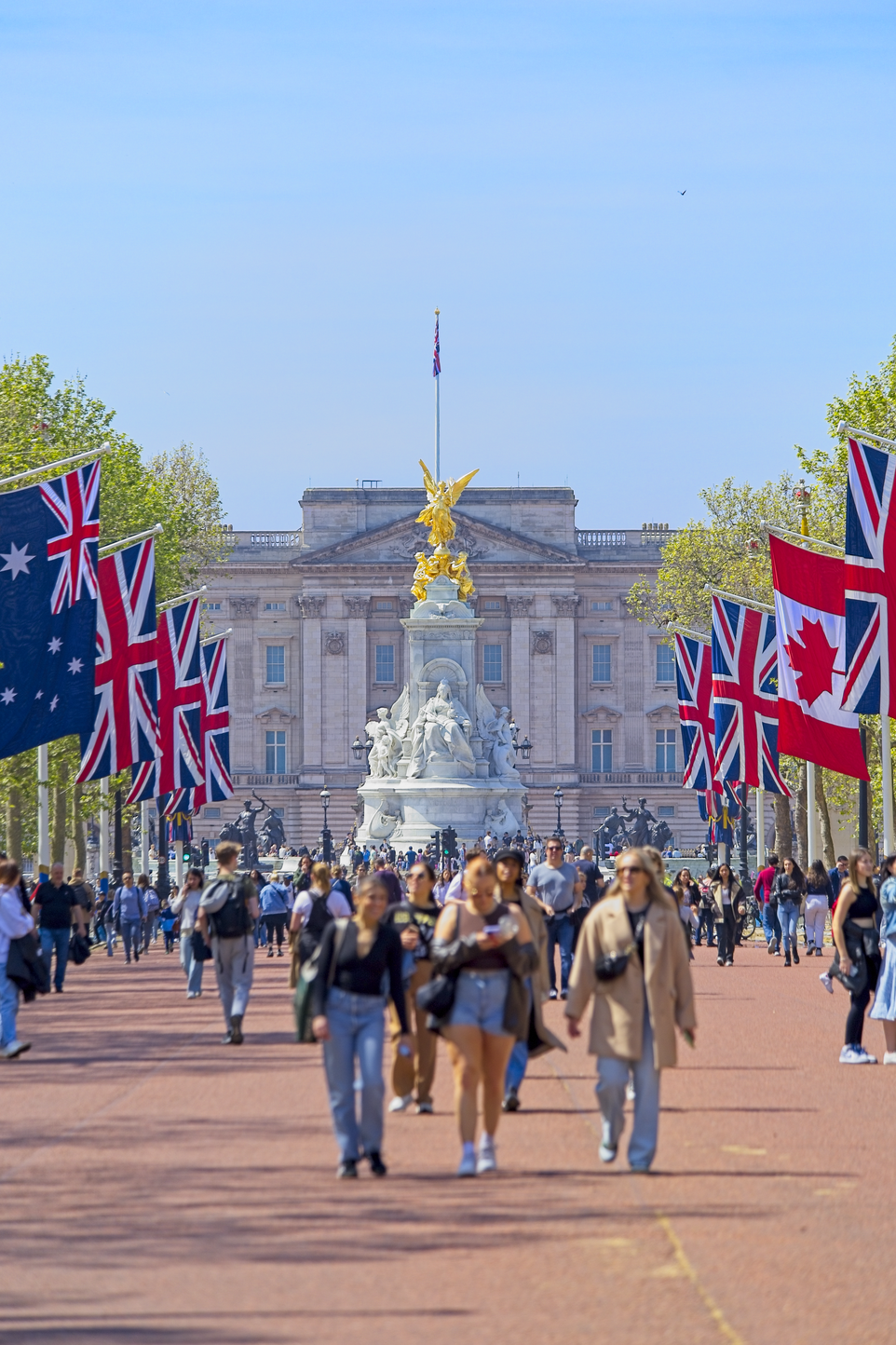 A bustling scene in a large, sunny plaza, featuring numerous international flags (including the Union Jack and Canadian flags). People are casually strolling across the foreground toward a grand, classical building that features a central monument.