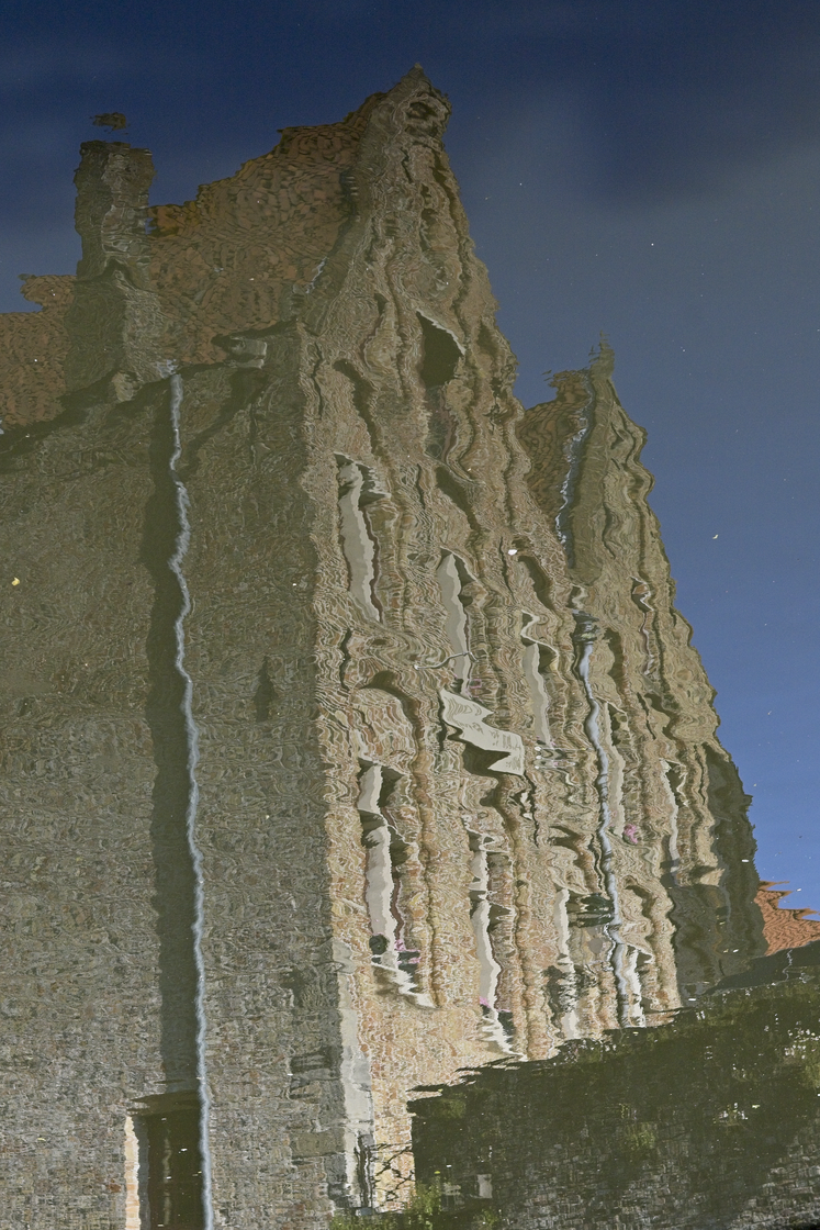 A distorted, rippled reflection of an old stone tower or castle-like building captured in still water. The wavy water surface creates an abstract, undulating pattern that warps the structure's vertical lines and architectural details, with vertical streaks and shimmering highlights. A clear blue sky is visible in the background, and darker reflections of surrounding walls and vegetation appear at the bottom of the frame.
