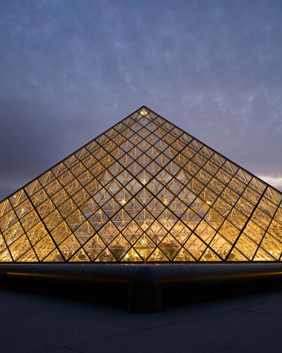 An architectural photograph capturing the iconic glass Louvre Pyramid at dusk. The structure is brightly illuminated, creating a grid-like pattern of light that contrasts dramatically with the moody, overcast twilight sky above.