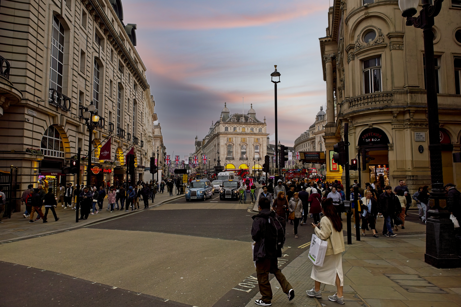 A bustling urban scene in a historic European city, likely London, captured during twilight. Grand, classical architecture lines the wide street, which is busy with pedestrians enjoying the late afternoon atmosphere.