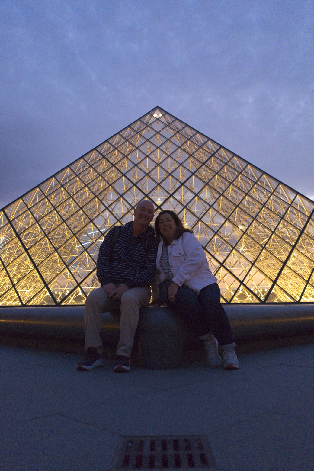 A couple is enjoying a twilight view in front of a massive, illuminated geometric pyramid structure. The warm, golden glow of the architecture contrasts with the cool blue tones of the dusk sky.