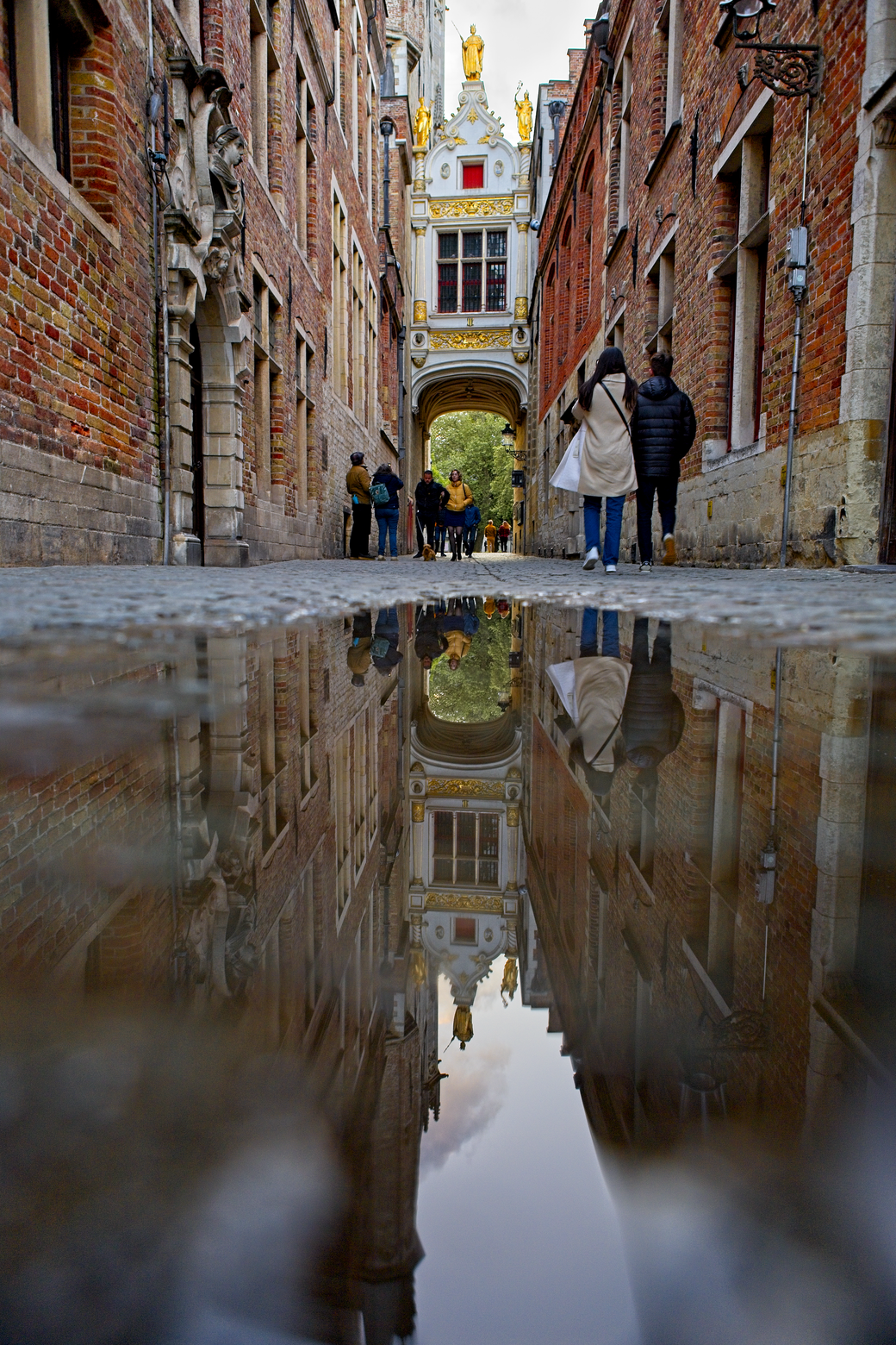 A dramatic street scene featuring historic, brick-lined architecture, viewed through a highly reflective wet surface. The image captures a strong sense of symmetry, with the surrounding buildings, pedestrians, and an ornate central gateway mirrored perfectly on the foreground puddle, giving the street a surreal, reflective quality.