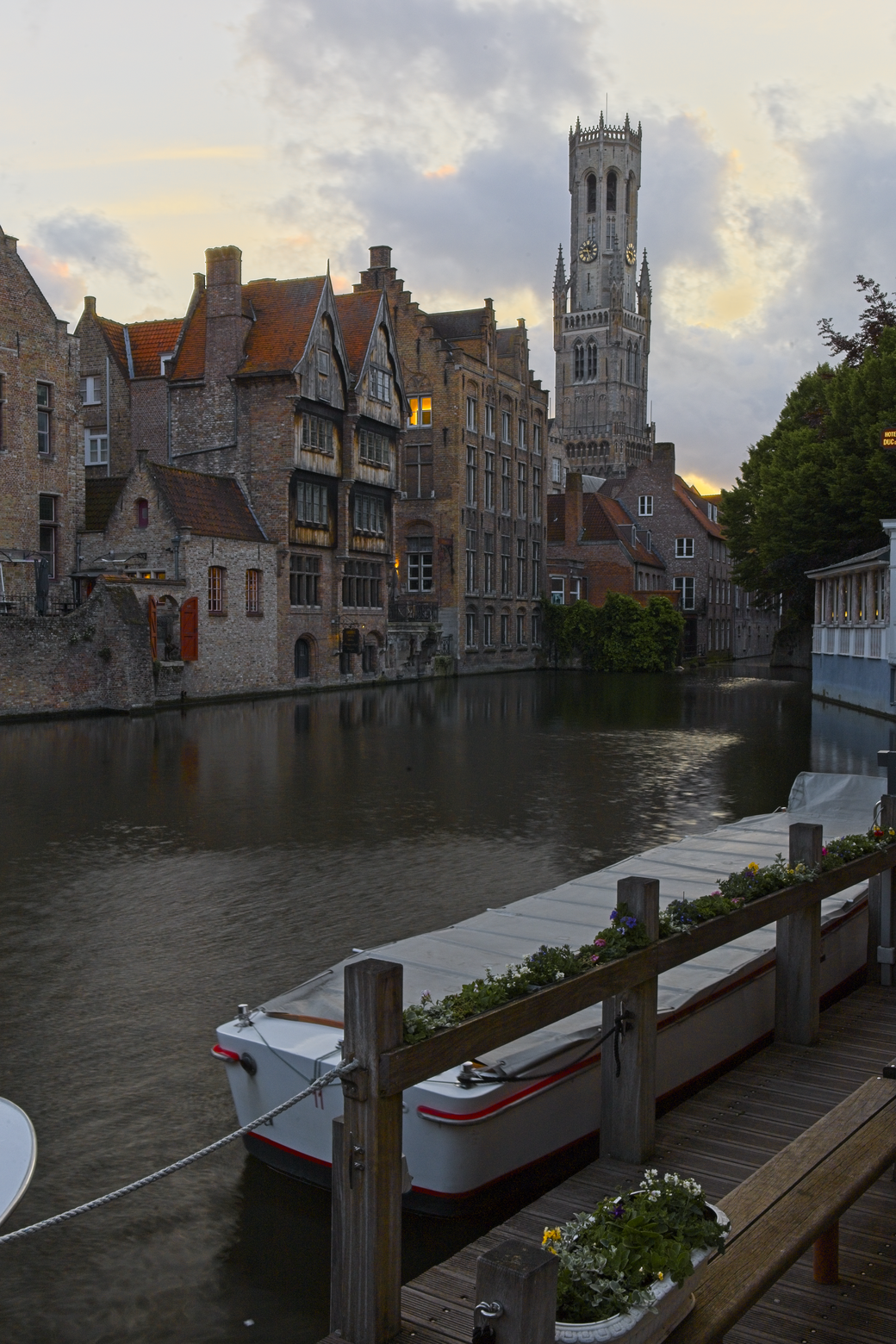 A picturesque view of a historic European city canal, featuring traditional gabled buildings lining the waterway. The scene is dominated by a large, prominent bell tower (belfry) on the right. In the foreground, a moored canal boat rests against a wooden dock, all framed by a dramatic, cloudy sky.