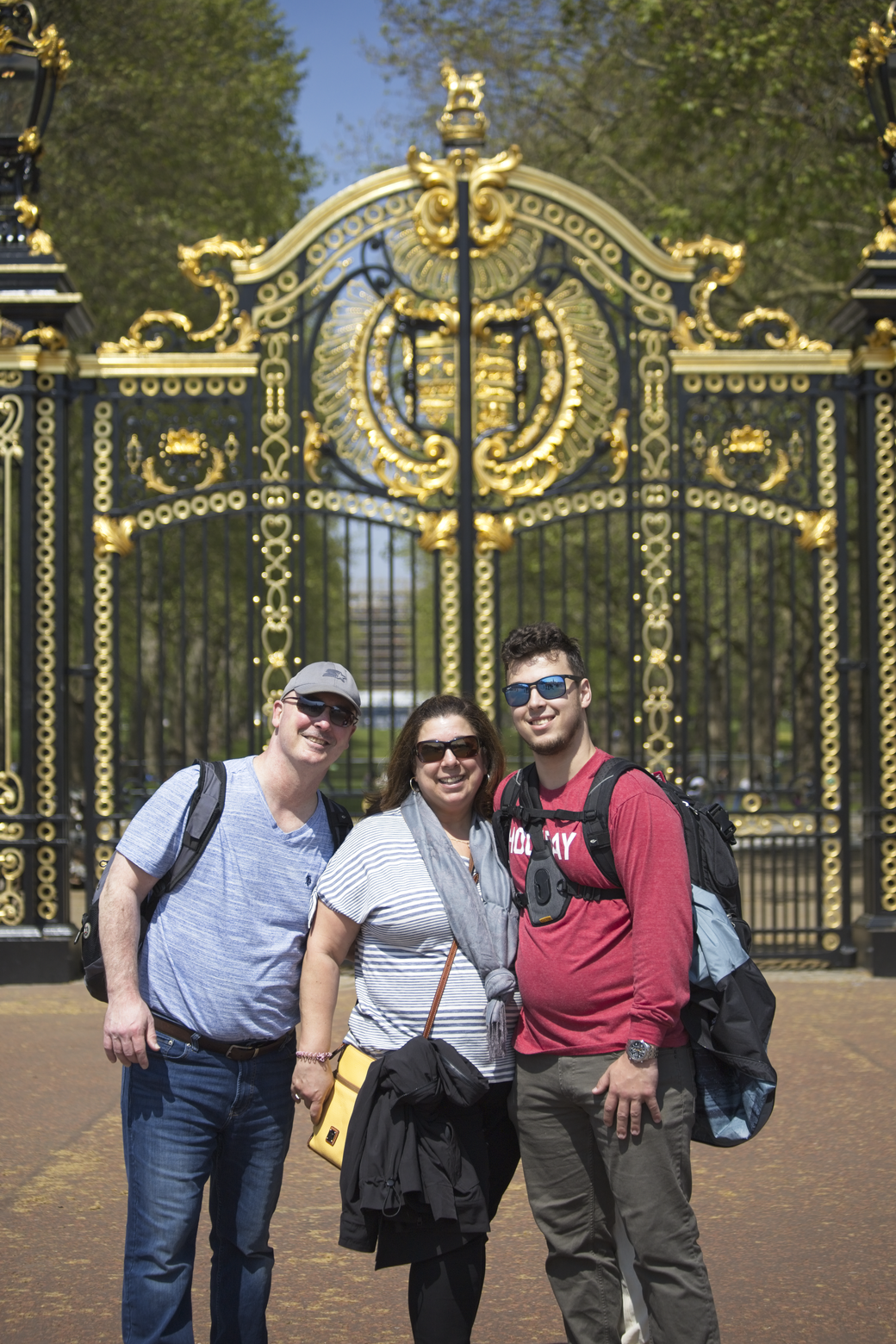 A group of three friends (two men and one woman) posing for a photo in front of an ornate, black and gold wrought-iron gate, suggesting a visit to a historic park or location.