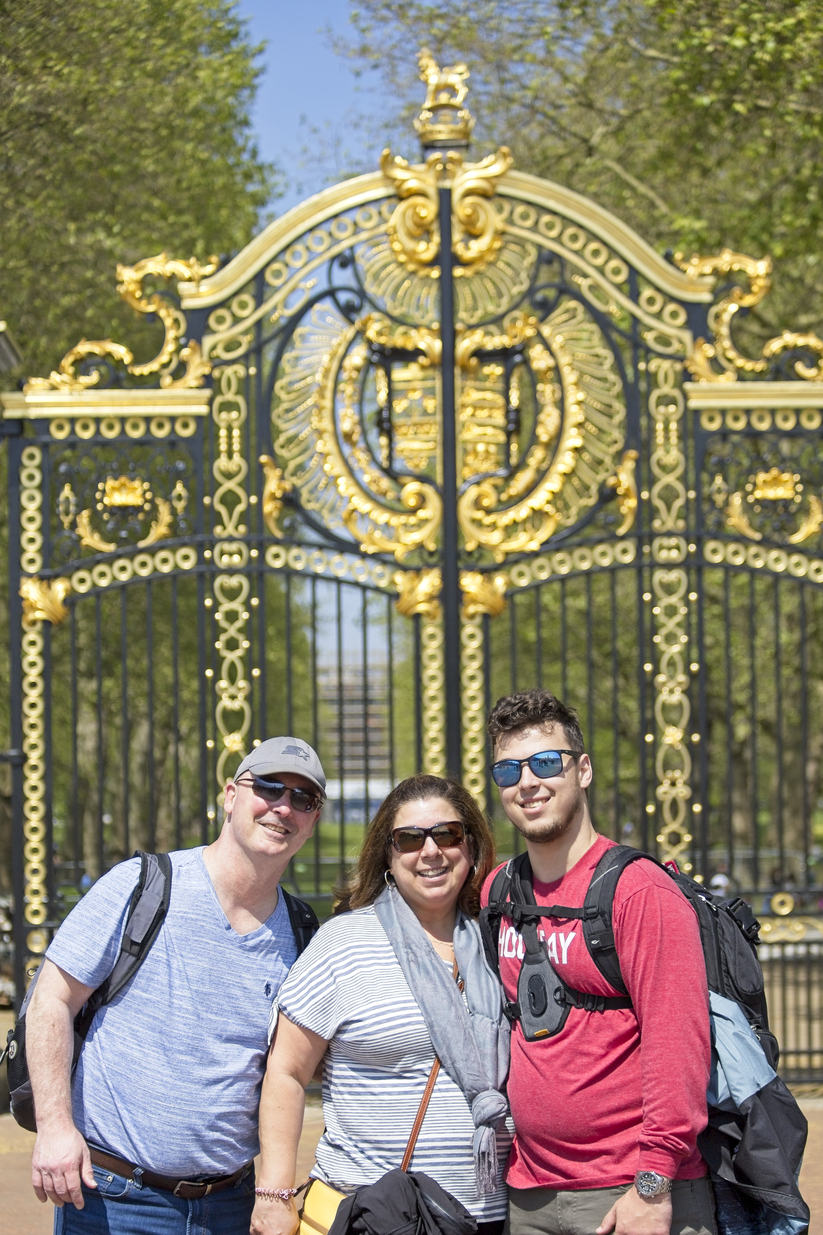 A group of four friends is posing in a park, standing in front of a large, ornate wrought-iron gate detailed with gold accents.