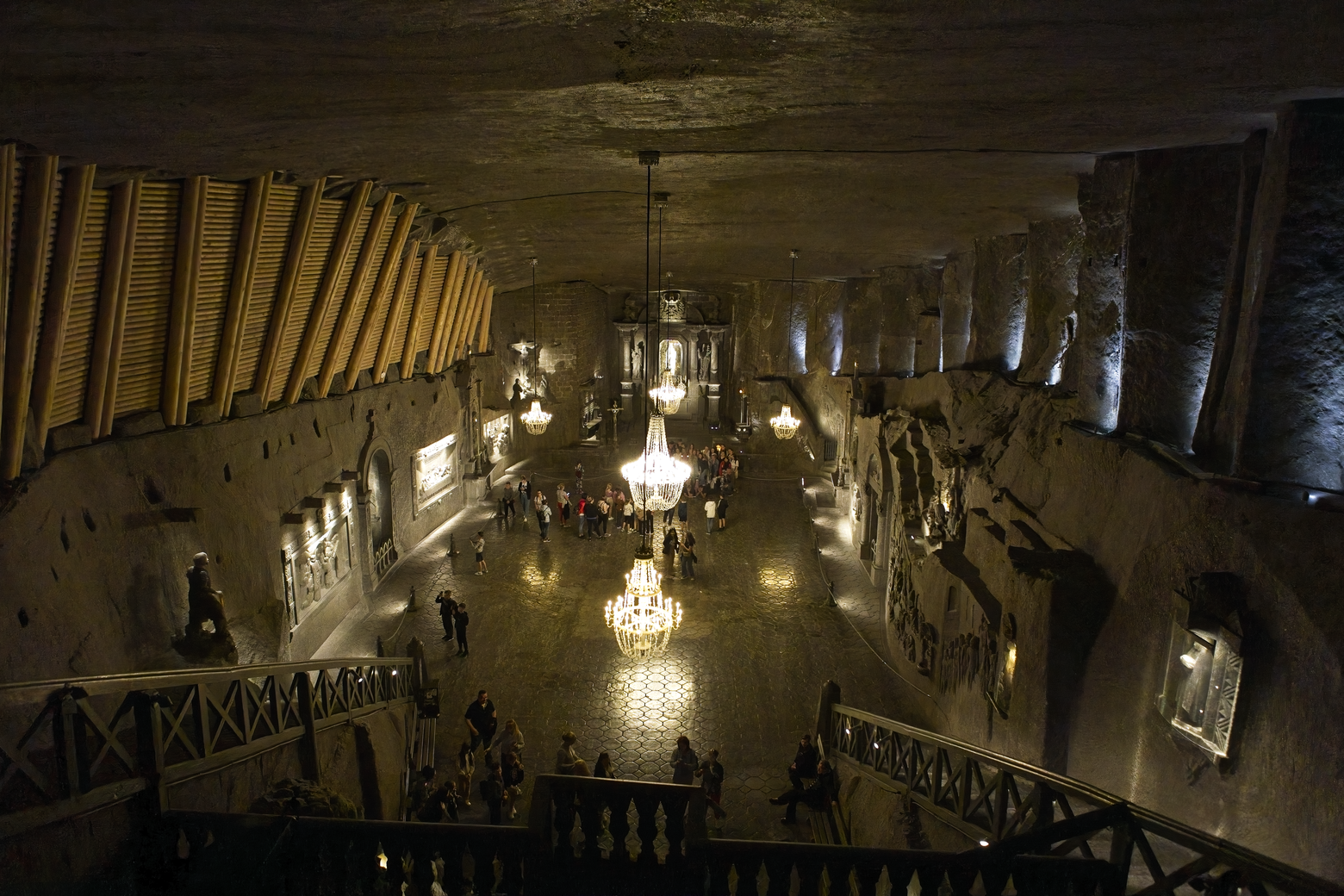 The image captures a dramatic view of a large, vaulted indoor public space, reminiscent of a historic market hall or underground arcade. The area is illuminated by multiple ornate chandeliers and ambient lighting, drawing the eye toward a central gathering point where people are visible. The architecture features high, curved stone walls and a substantial ceiling, creating a moody, yet atmospheric, setting.