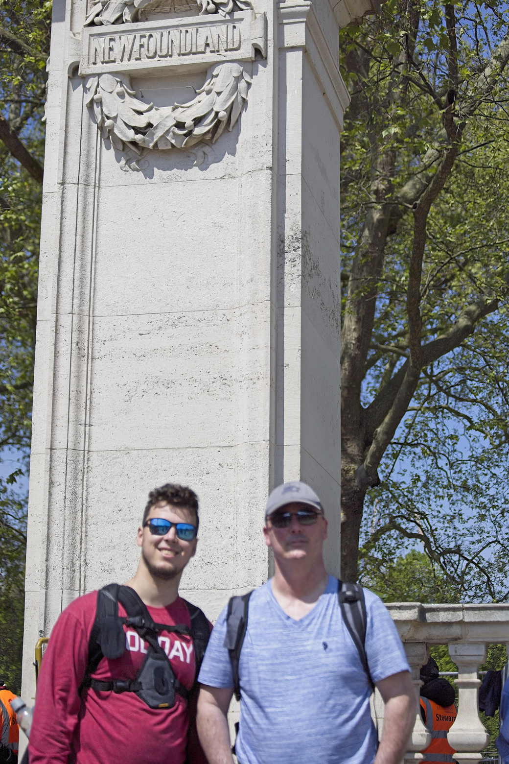 Two men are standing outside, posing in front of a large, white monument that features the inscription 'NEWFOUNDLAND.' The setting is outdoors, characterized by lush green trees and clear weather, suggesting a historical landmark or memorial.