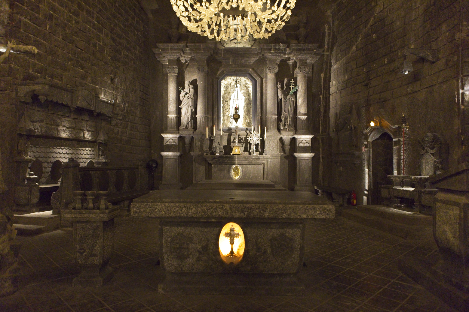 A dramatic, atmospheric photograph of a dimly lit church sanctuary. The scene is dominated by a large, ornate chandelier and features a central altar niche flanked by statues and religious iconography. The overall ambiance is solemn, historical, and reverent, emphasizing the rough texture of the stone walls.