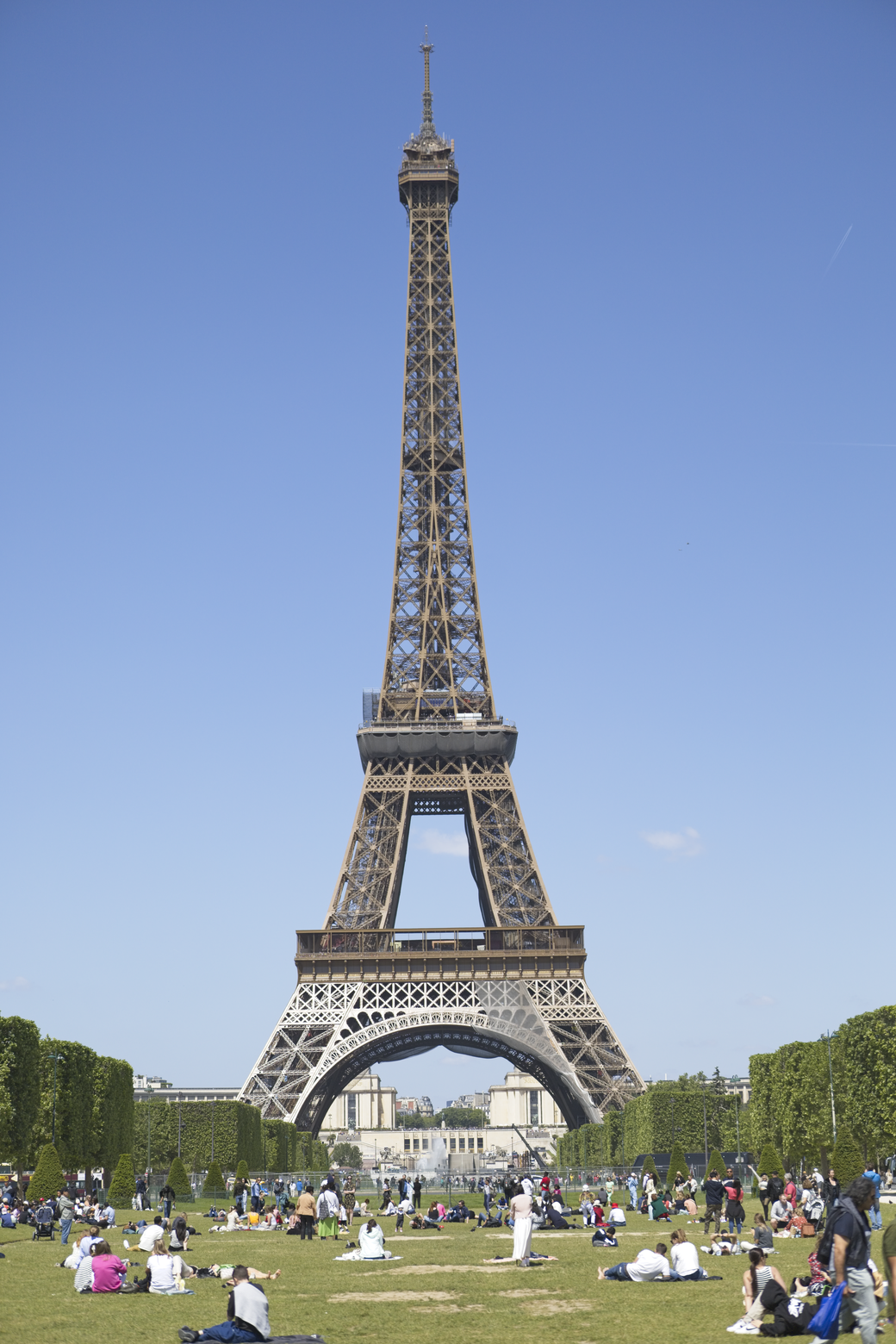 A panoramic view of the Eiffel Tower dominating the bright blue sky. In the foreground, people are gathered and relaxing on the expansive green lawn of the park, enjoying a clear, sunny day in Paris.