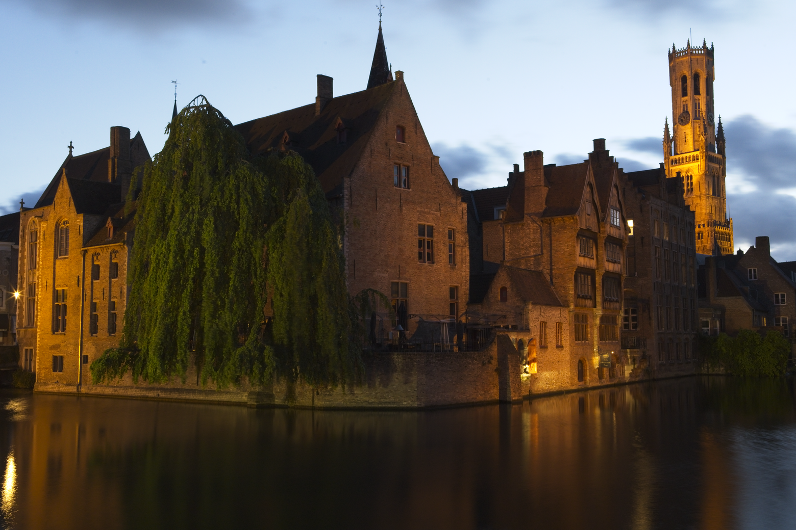 A serene twilight view of historic European canal architecture. Gabled buildings line the waterfront, featuring a prominent church tower on the right. A large, weeping tree dominates the foreground, while the calm canal water reflects the warm glow of the structures against the deep blue sky.