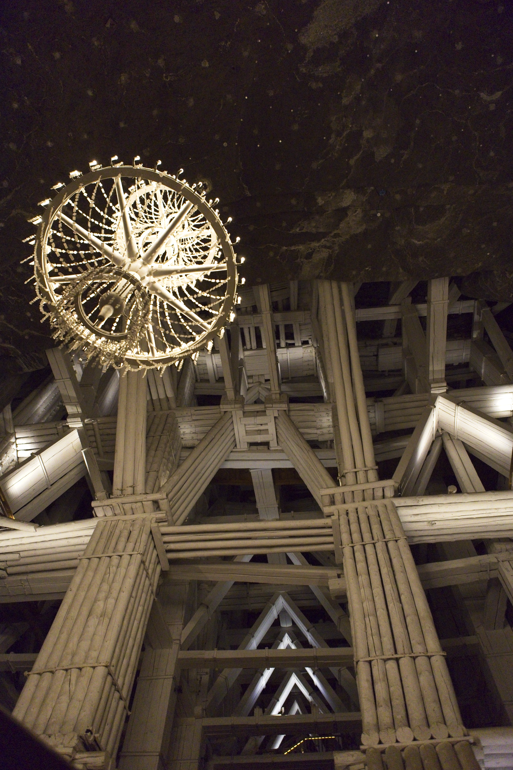 A dramatic, low-angle photograph capturing the immense interior of a large public structure, such as a train station. The scene is dominated by a gigantic, illuminated Ferris wheel suspended beneath a vast, dark ceiling. The architecture features massive, intersecting columns and structural trusses, emphasizing the overwhelming scale and industrial grandeur of the space.