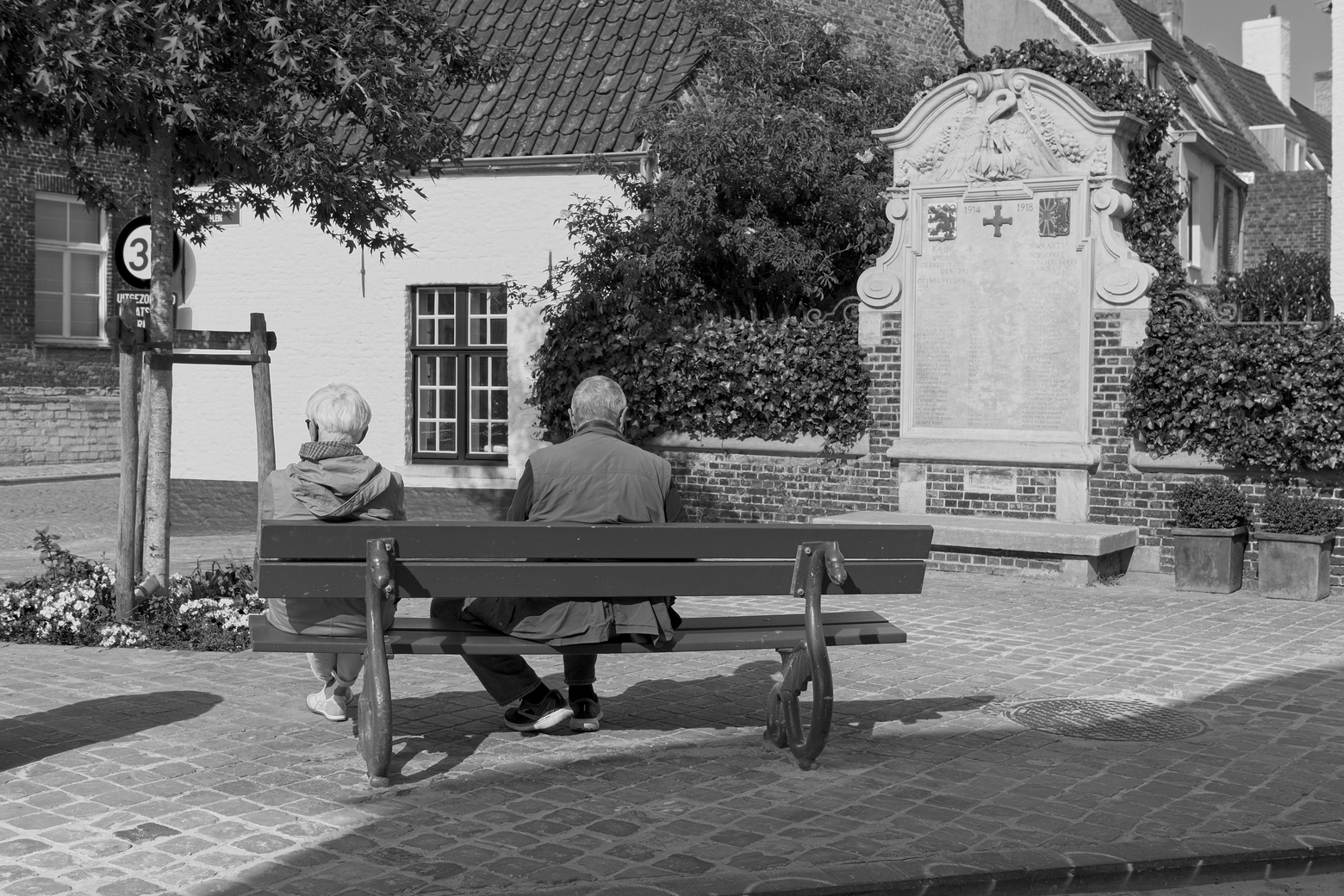 A black and white photograph of two elderly people sitting on a park bench with their backs to the camera, gazing at an ornate stone war memorial or monument. The scene is set in a charming European village square with cobblestone paving, surrounded by white and brick buildings, ivy-covered walls, a small tree, and potted plants.