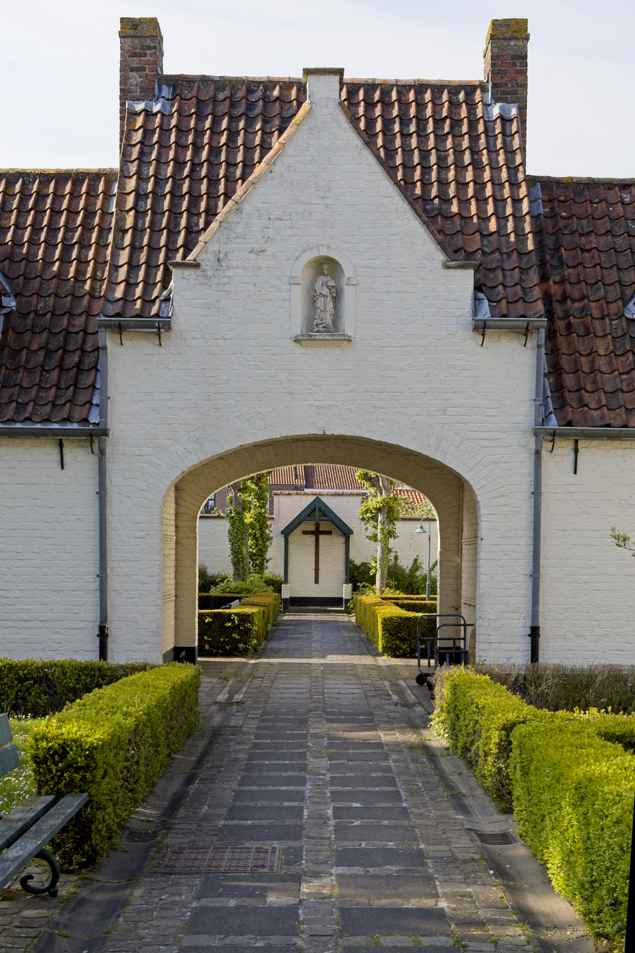 A view looking through a large, arched white entrance of a historic building, likely a church or monastery. The path leading through the arch is flanked by neatly trimmed green hedges, guiding the eye toward an inner courtyard visible in the background. The structure features a prominent tiled roof and a niche statue above the main gateway.