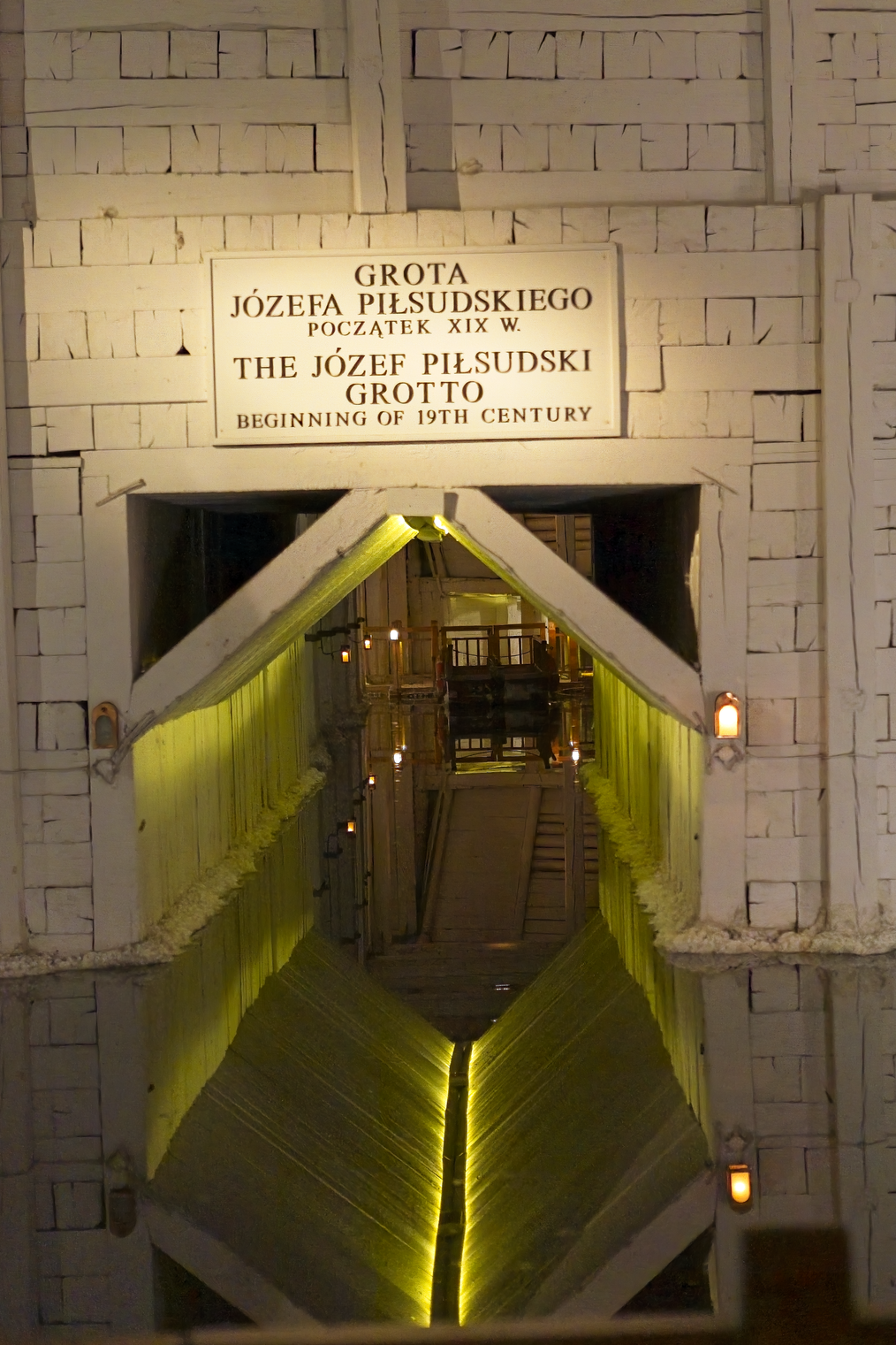The image captures the entrance to the Józef Piłsudski Grotto (Grotta Józefa Piłsudskiego), a memorial site. The entranceway is characterized by white brickwork and features multilingual signage identifying the grotto and placing its historical context at the beginning of the 19th century. The internal passageway is dramatically lit with warm, yellow lights, creating an immersive and historical atmosphere.
