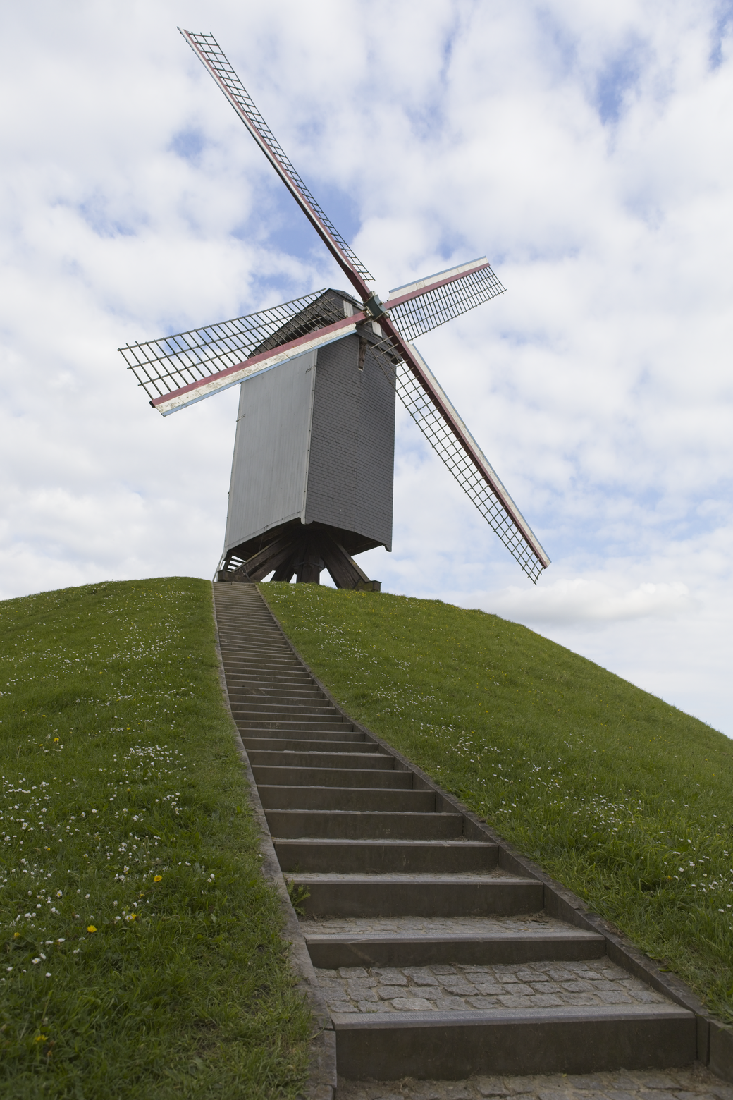 A scenic view featuring a traditional windmill positioned atop a grassy hill. A wide staircase leads the eye up the lush green slope toward the structure, which is framed beneath a partly cloudy blue sky.
