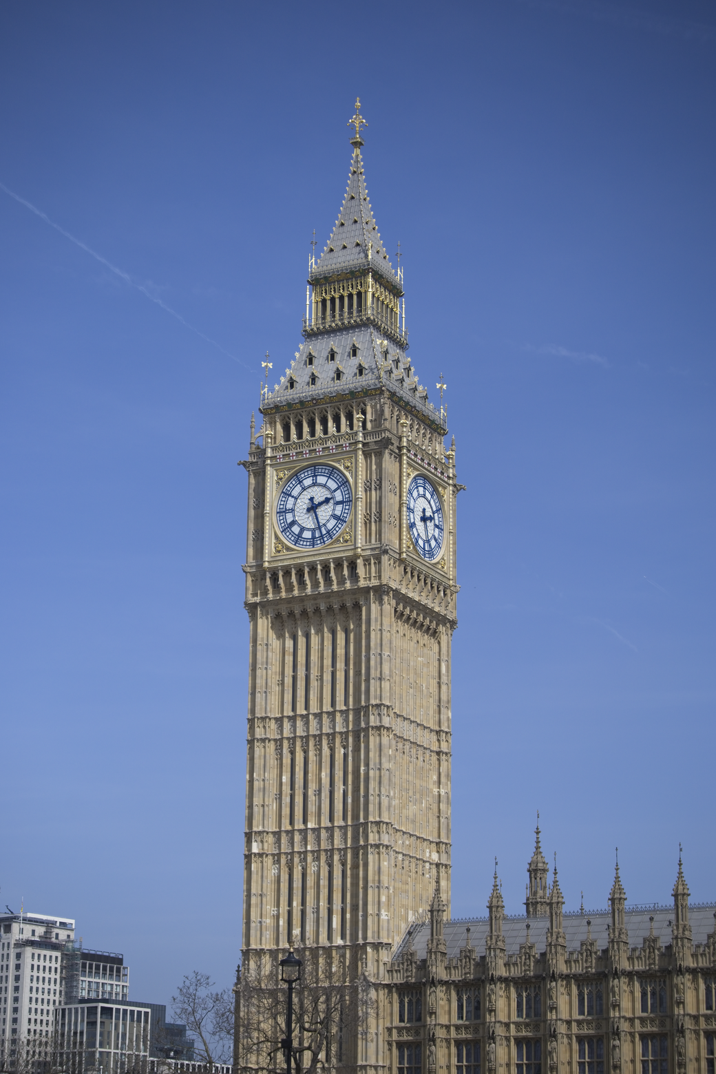 A view of the iconic Elizabeth Tower (Big Ben) dominating the center of the frame, set against a clear blue sky, with the complementary architecture of the Palace of Westminster visible in the foreground.