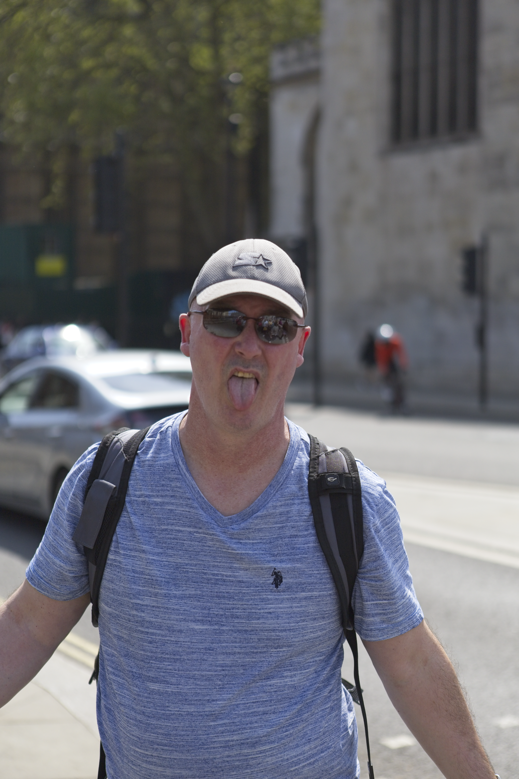 A man wearing a t-shirt, sunglasses, and a backpack stands on an urban sidewalk, playfully sticking his tongue out towards the camera. The background features a city street with buildings and parked cars visible.