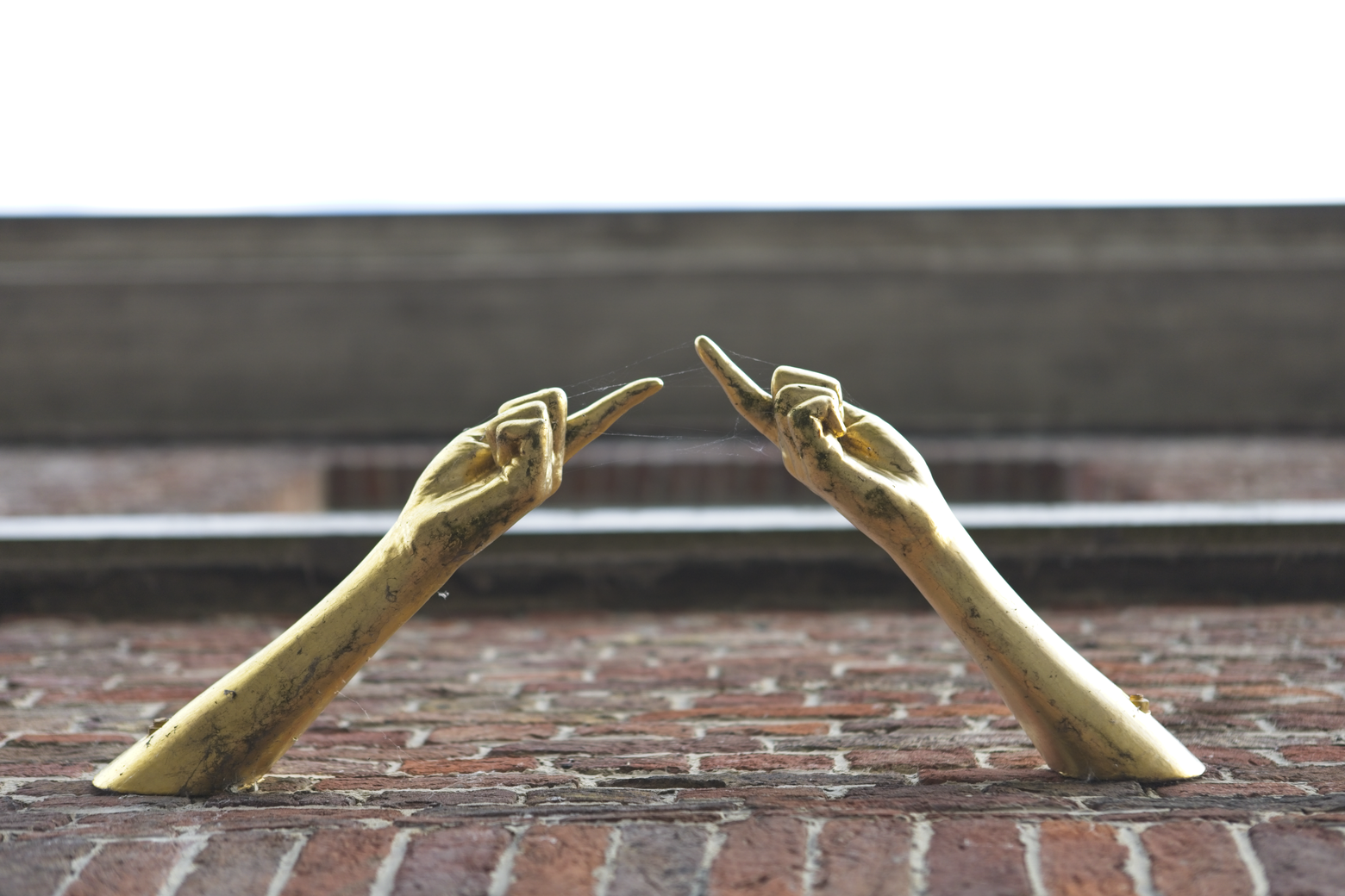 Two stylized, golden metallic arms are positioned pointing inward towards each other, resting upon a textured reddish-brown brick surface. The background is minimalist, consisting of a dark horizontal ledge and a bright white sky, focusing attention on the sculptural elements.