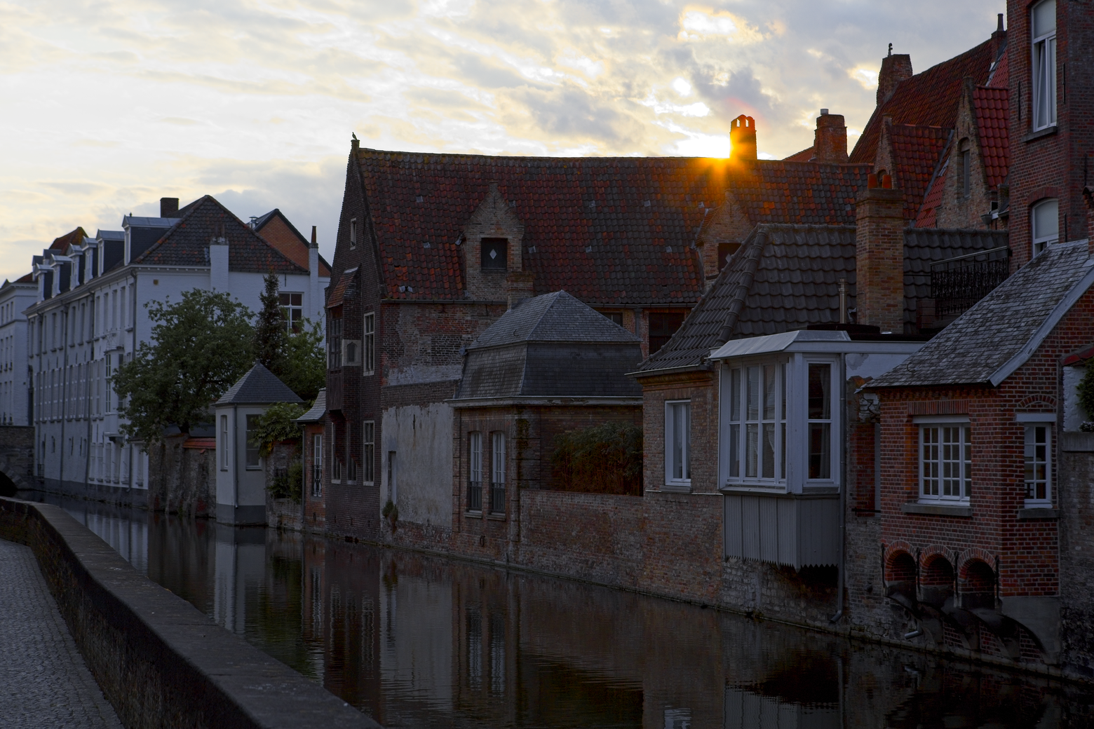 A picturesque canal scene in an old European city, featuring rows of historic brick buildings with tiled roofs. The soft light of sunrise or sunset shines across the waterway, creating dramatic reflections on the calm water and highlighting the classic architecture.