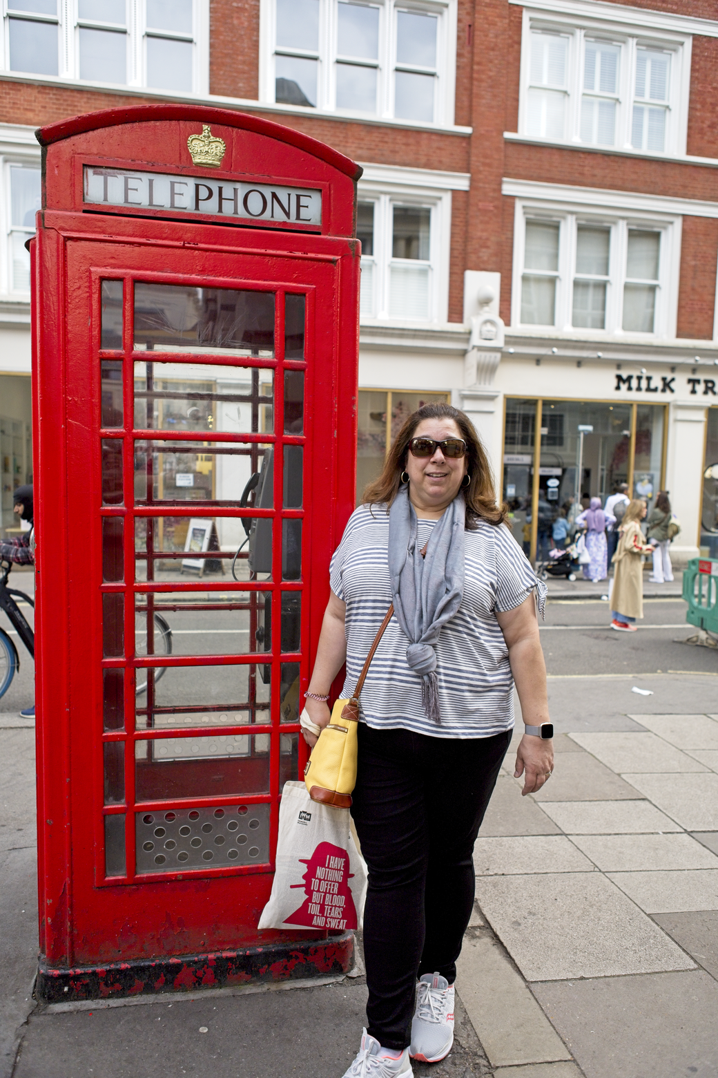 A woman standing on a city sidewalk next to a classic red British telephone box. She is dressed casually and is posing in front of a large, multi-story building that features storefronts.