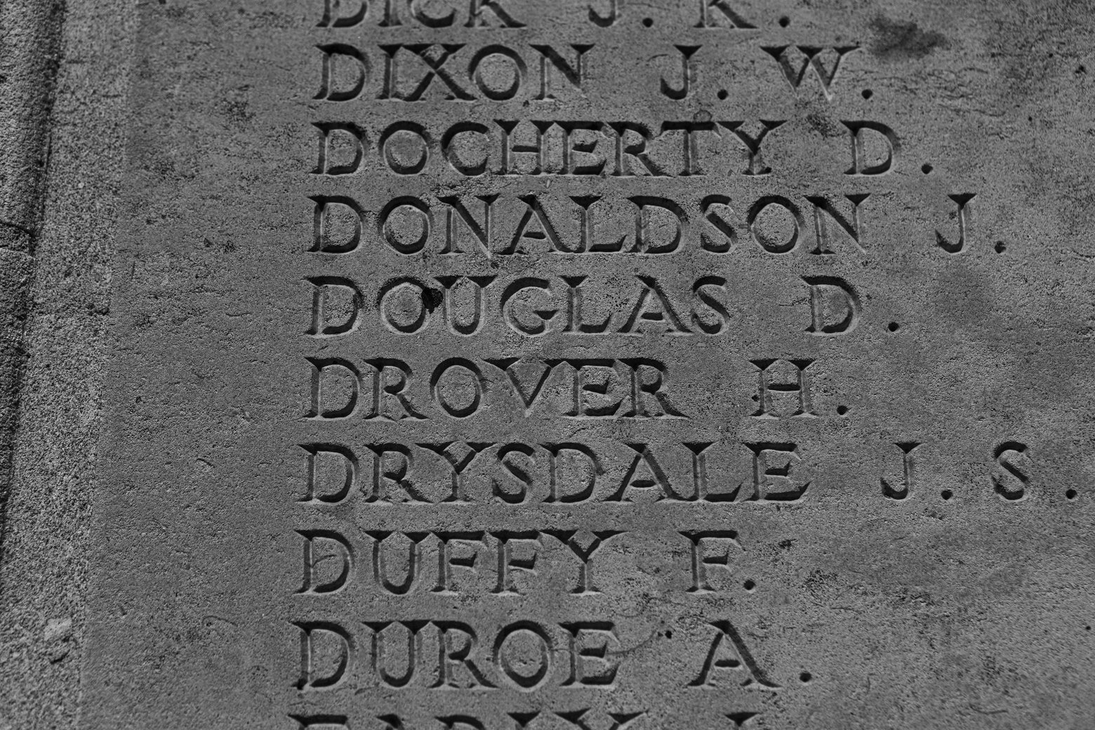 A black-and-white close-up of a weathered stone memorial or headstone featuring engraved uppercase names arranged alphabetically. Visible inscriptions include DICK, DIXON J.W., DOCHERTY D., DONALDSON J., DOUGLAS D., DROVER H., DRYSDALE J.S., DUFFY F., and DUROE A., with portions of names cut off at the top and bottom edges. The stone surface shows a rough, aged texture with chiseled lettering and small weathering marks.