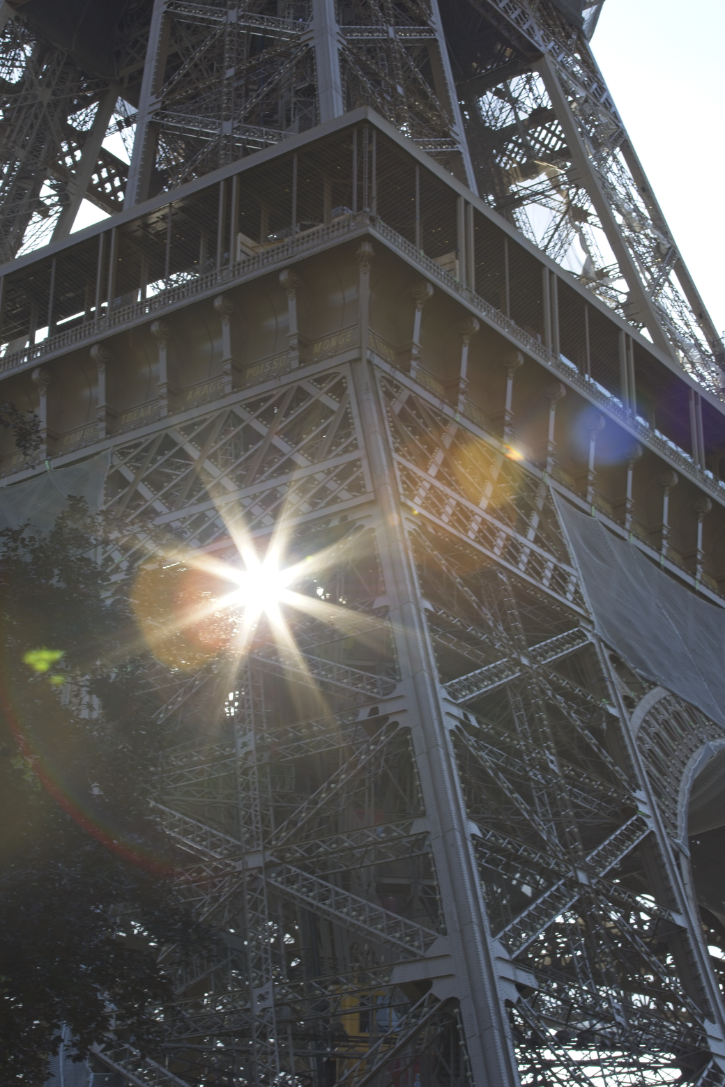 A dramatic, low-angle photograph of the Eiffel Tower, featuring strong backlighting that creates prominent sun flares across its intricate iron lattice structure. The perspective emphasizes the immense height and grand scale of the iconic Parisian monument.
