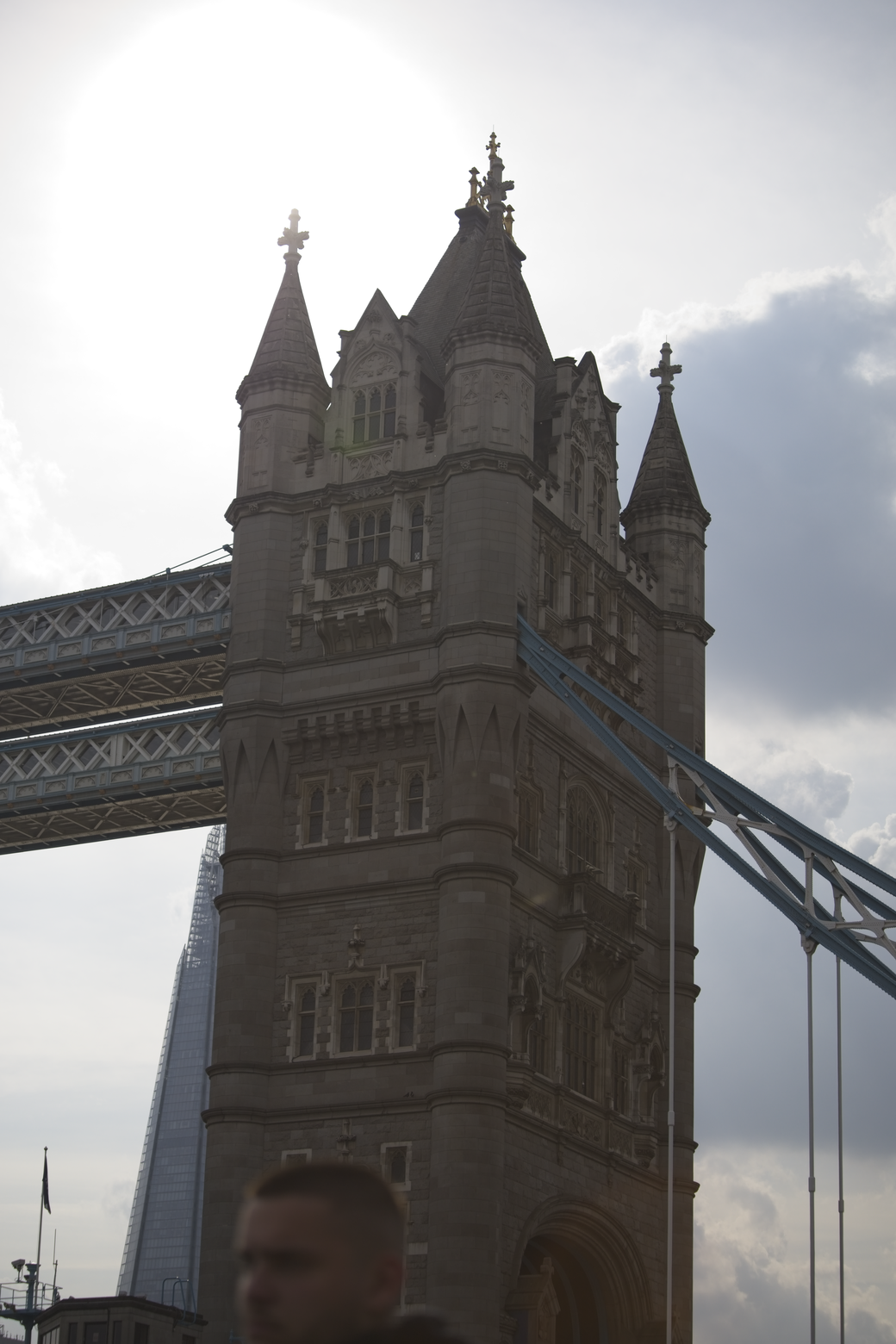 The image features a large, historic stone tower with ornate turrets and detailed masonry, suggesting grand European architecture. The monument is situated adjacent to a massive metal truss bridge structure. The scene is captured outdoors under a bright, cloudy sky, giving the architectural elements an imposing, high-contrast silhouette.