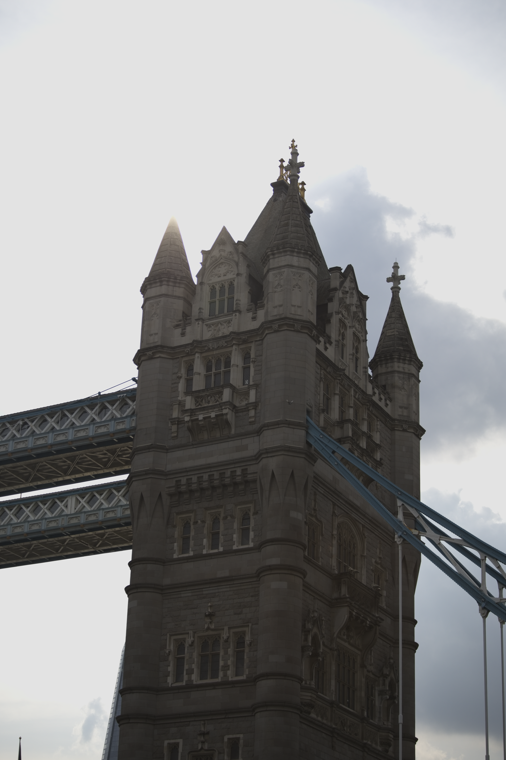 A low-angle view of a large, historical stone building featuring turrets and ornate architecture, set against an overcast sky, with a large truss bridge running in the foreground.