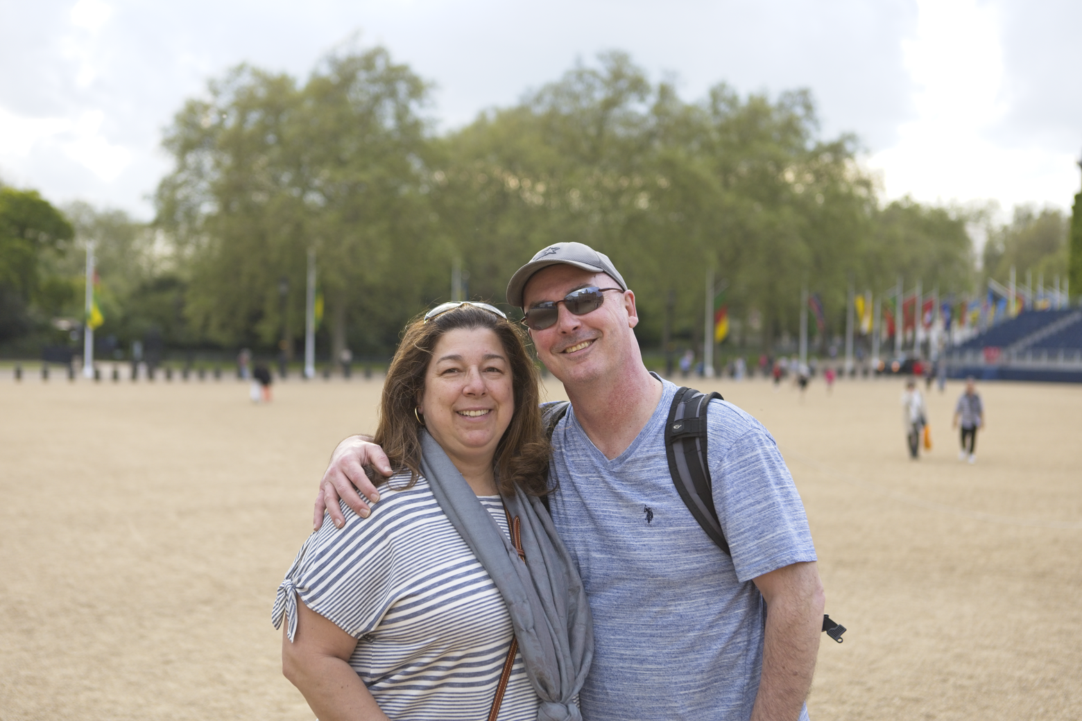 A cheerful photograph of a couple posing for a picture in a large, open outdoor park setting. The woman is leaning into the man, and both are smiling while standing on a wide, grassy field with trees visible in the background.
