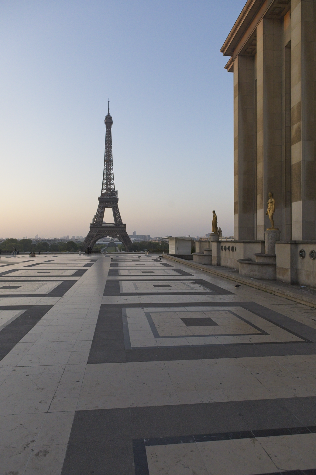 A view of the Eiffel Tower seen across a large, patterned plaza, framed on the right by the massive facade of a classical building, all set beneath a pale sky.