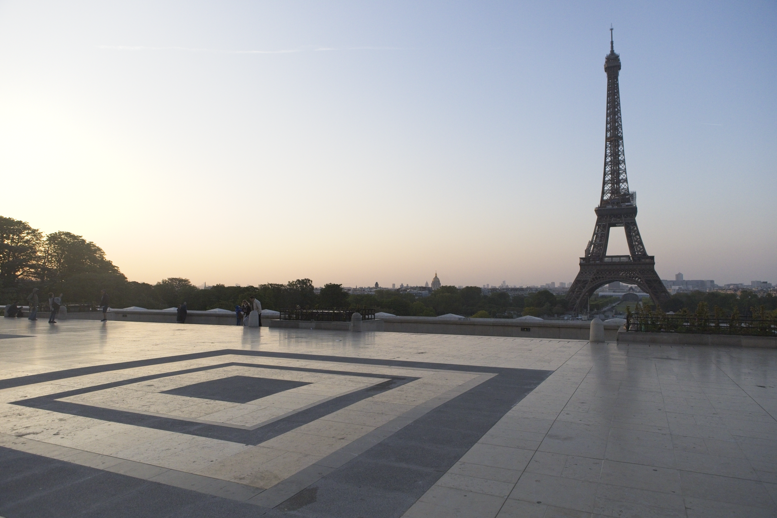 A wide-angle view featuring the Eiffel Tower set against a bright, soft-lit sky, likely near sunset. In the foreground, there is a patterned stone plaza, creating a grand and open perspective that frames the iconic Parisian landmark.