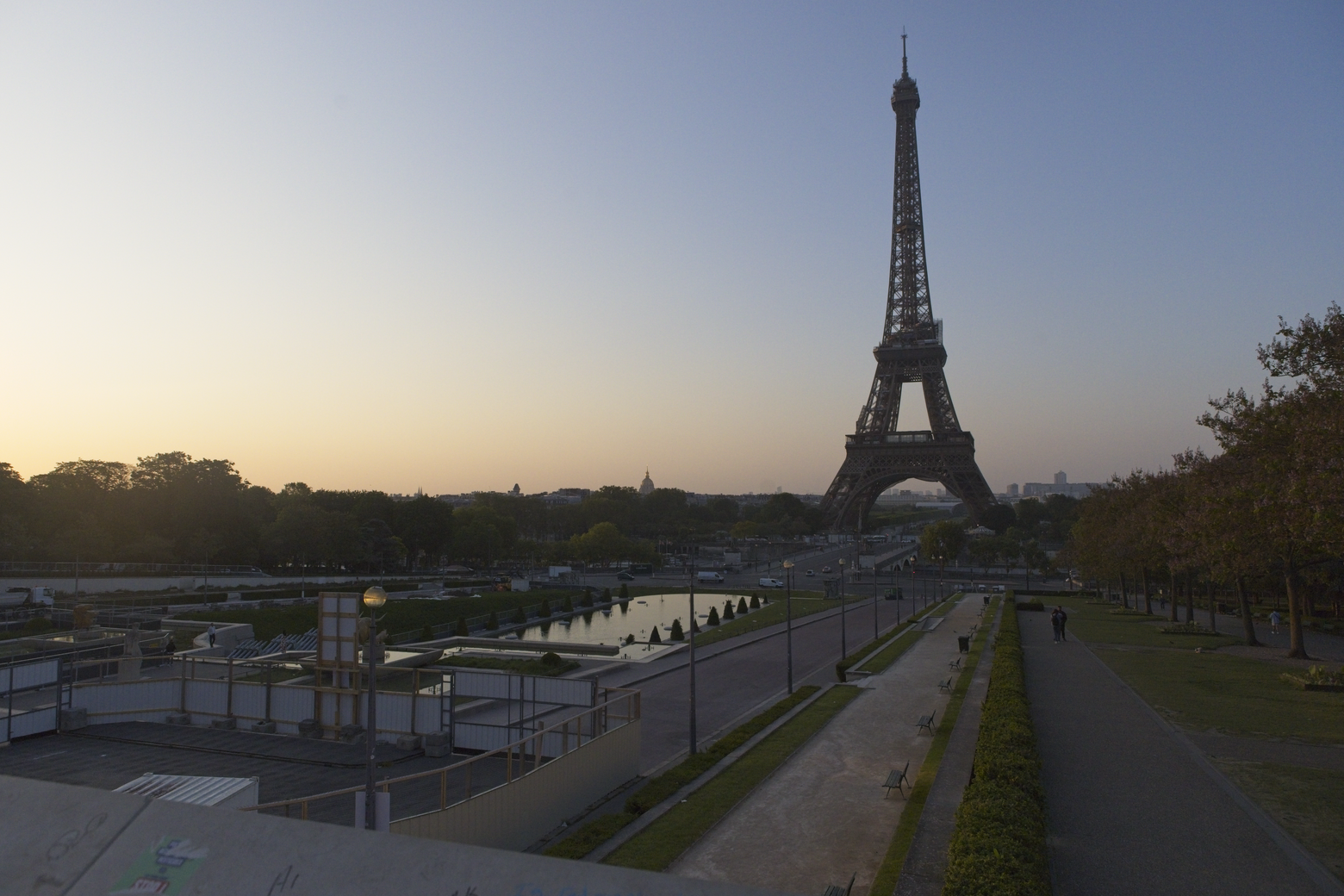 A panoramic view of the Eiffel Tower rising against a soft, twilight sky. The foreground features a wide, landscaped park area with pathways, foliage, and water features, capturing the Parisian landmark in a tranquil, daytime/twilight setting.