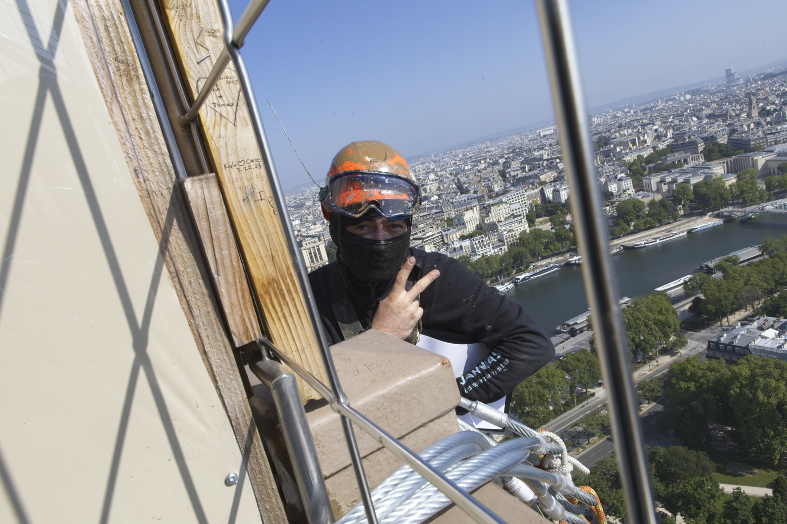 A person stands on an elevated viewing platform, looking out at a vast, sprawling cityscape. The foreground is dominated by metal railings and architectural structures, framing the expansive view of a densely built European city crisscrossed by rivers under a clear blue sky.